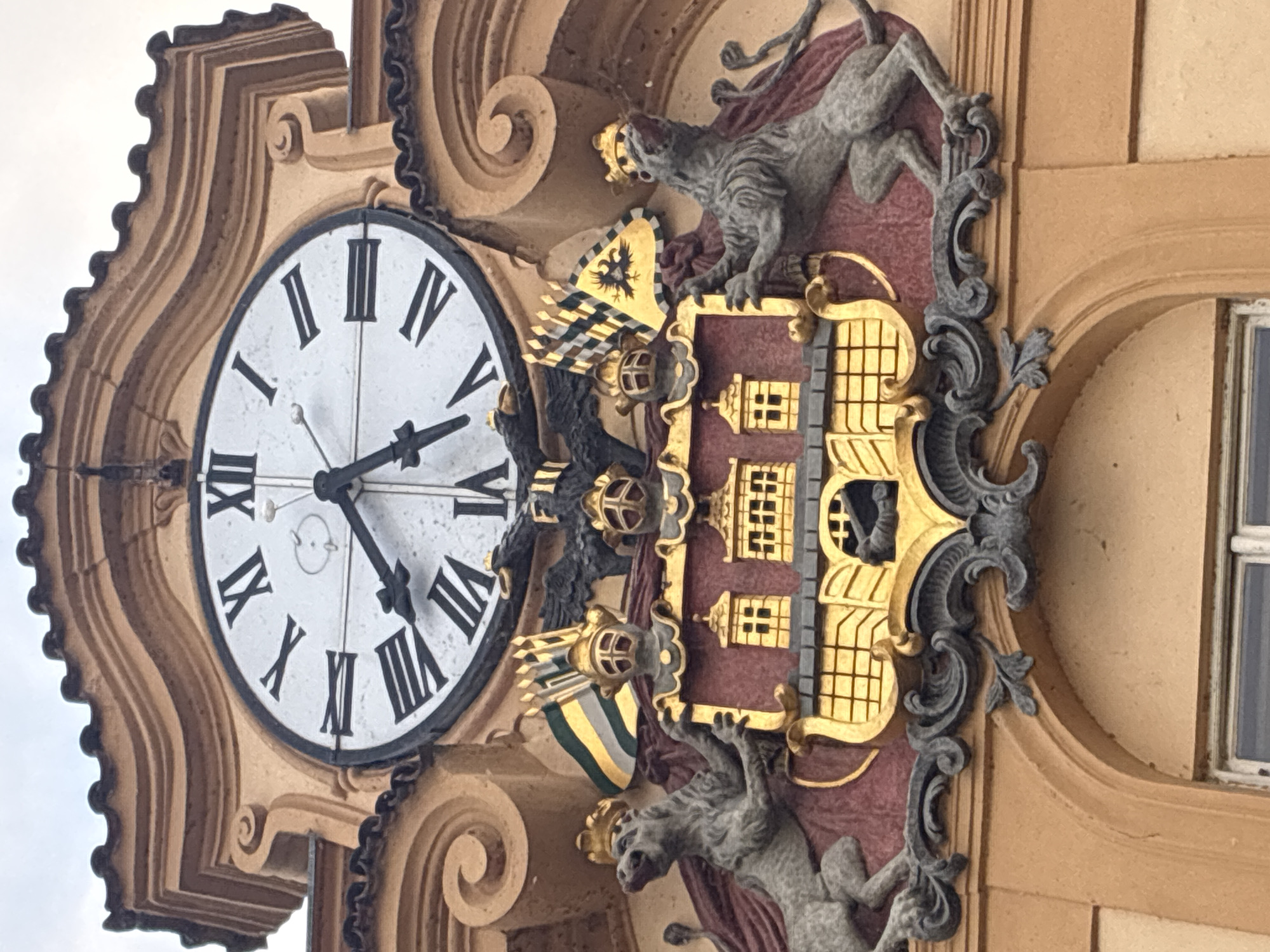The image shows a close-up view of a large clock on the facade of an ornate building. The clock has Roman numerals and ornate black hands, with the time appearing to be approximately 10:07. Below the clock is a detailed coat of arms featuring two crowned lions rampant, supporting a shield. The shield is emblazoned with a golden castle or town with multiple windows, set against a dark red background. Above the shield are two eagle heads. Behind the shield are various banners and flags, some bearing what appear to be heraldic symbols. The architecture of the building surrounding the clock and coat of arms is elaborate, with carved decorative elements in a golden-brown hue. The overall impression is one of historical grandeur. This is the Astronomical Clock (Orloj) located in Old Town Square in Prague, Czechia.