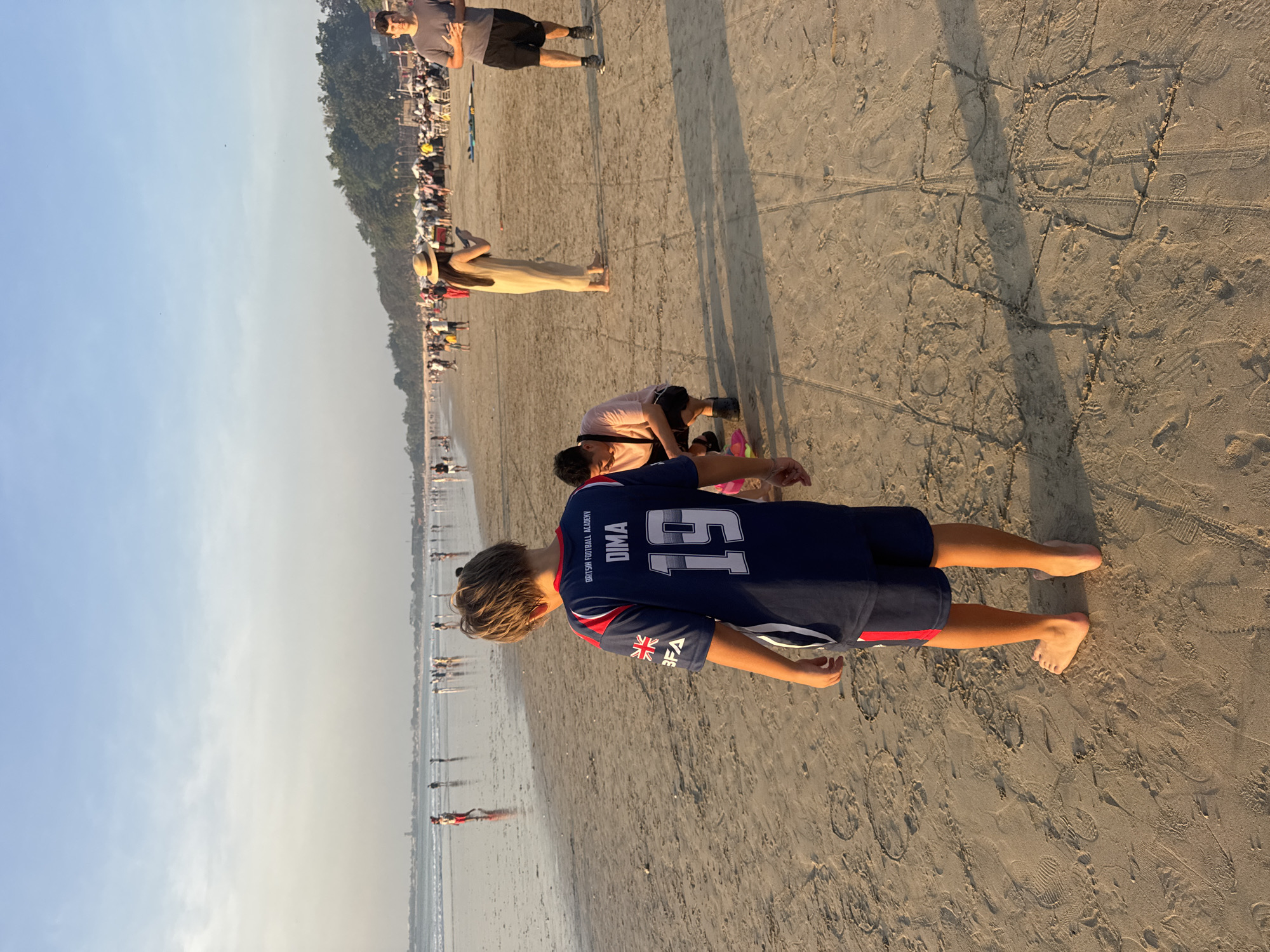 In Kuta Selatan, Indonesia, this image captures a beach scene at sunset with several individuals present. In the foreground, a young boy with blonde hair faces away from the camera, wearing a blue and red football jersey with "DIMA 19" and "BRITISH FOOTBALL ACADEMY" printed on the back, along with a Union Jack flag emblem and "BFA." He is barefoot. To his right, another person is partially visible, wearing a pink t-shirt and black shorts. Further back on the beach, a woman in a tan dress and straw hat holds a phone, and a man in a grey t-shirt and black shorts stands. Other people are scattered along the shoreline and in the background, with tents and structures suggesting a market or gathering area on the left side of the image. The sand is imprinted with footprints and drawn shapes. The sea is calm with gentle waves, and the sky is clear with a soft gradient of blue. Trees and hills are visible in the far distance.