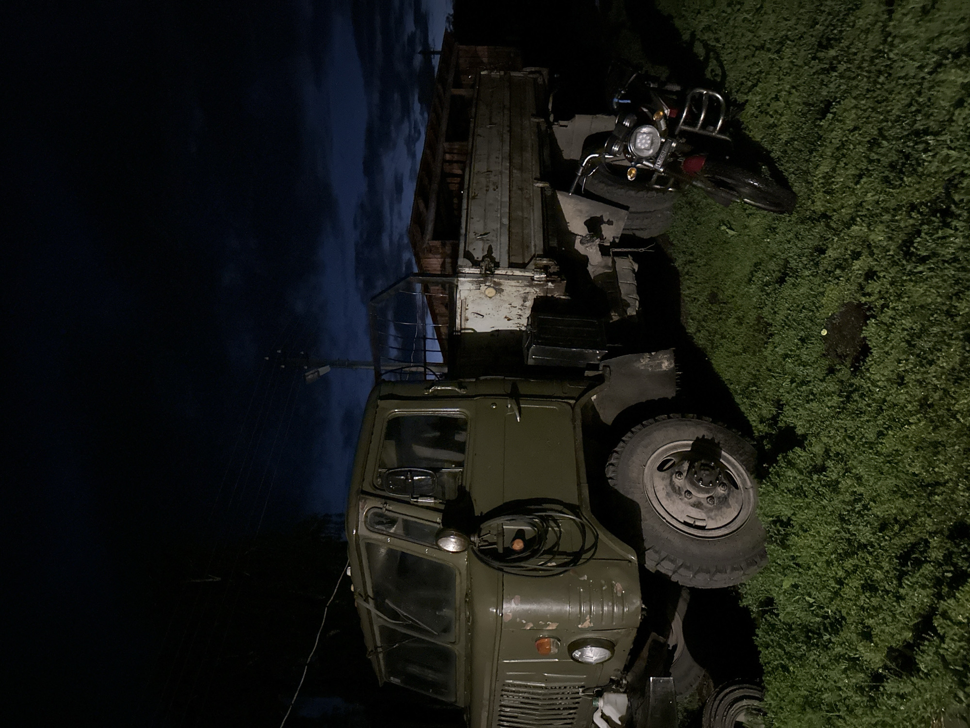 A dark, overcast night scene features a weathered, olive-green military-style truck parked on a grassy area. The truck has a large, multi-part windshield and a prominent grille. A coiled black hose hangs from a bracket on the driver's side. The truck's bed is elevated and appears to be made of rusted metal with wooden slats along the top edge. To the right of the truck, a small, dark-colored motorcycle with a bright LED headlight is parked. The background shows a dark, cloudy sky and a building structure with a partially visible roof. The overall lighting is dim, suggesting artificial light sources are limited or distant.