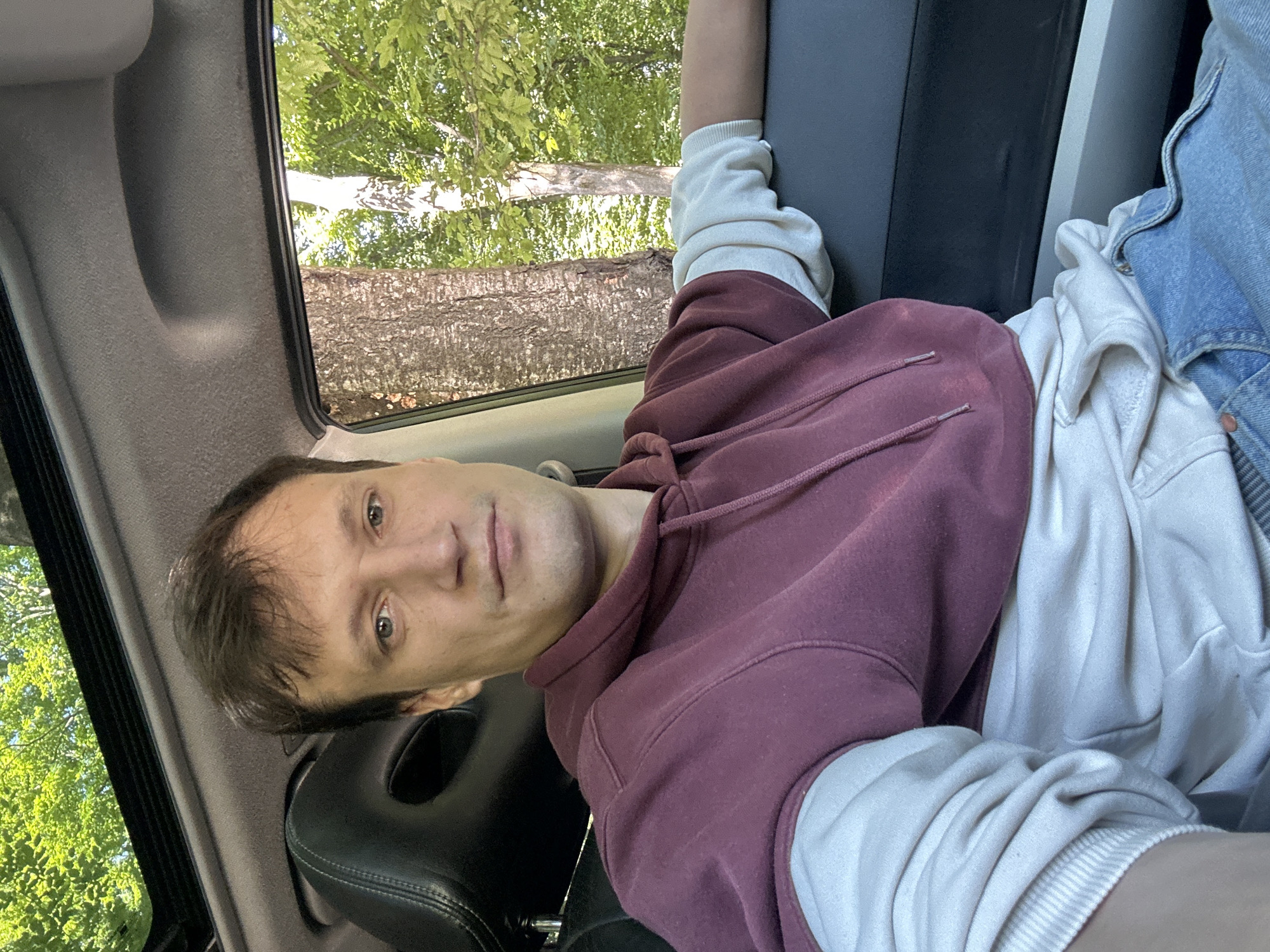 A man is sitting in the driver's seat of a car, looking at the camera. He has brown hair, blue eyes, and is wearing a maroon hoodie over a light-colored sweatshirt and blue jeans. The car's sunroof is open, revealing green trees and blue sky. The scene appears to be in a wooded area outside of Shatava, Ukraine, indicated by the trees and natural light.