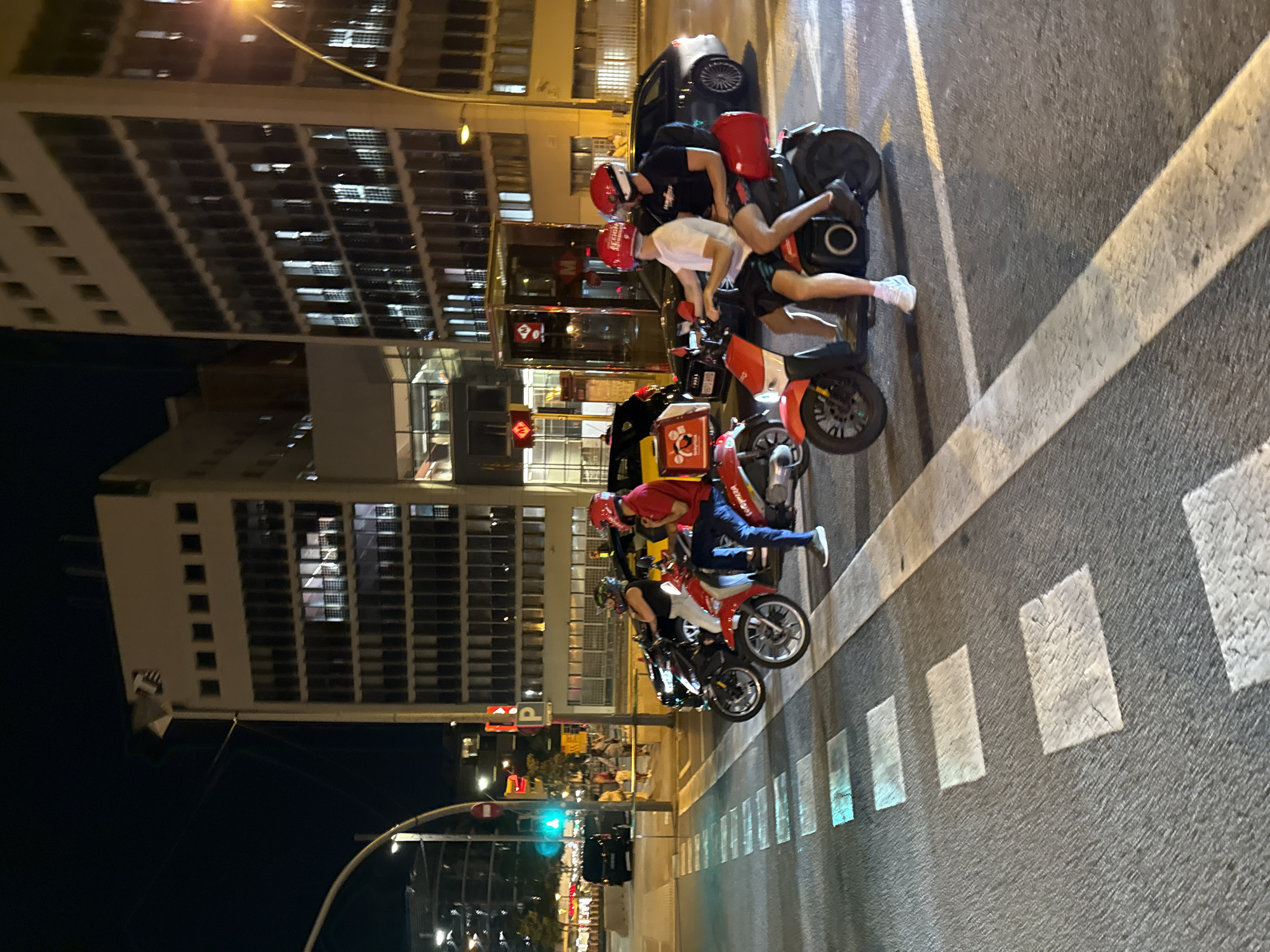 The image shows a nighttime street scene in Barcelona, Spain, near a Metro station. Several people on scooters are waiting at a crosswalk. Two of the scooters are red and appear to be delivery vehicles, one with a person in a red shirt and blue jeans and another with two people wearing red helmets. Another scooter is black, with a person wearing a black tank top and white shorts. Behind them, several cars are visible, including a black van and a silver Audi. The background features modern, multi-story buildings with numerous windows, some of which are lit. Streetlights and traffic signals are also present, with a green traffic light visible. The street is marked with white lines and rectangular pedestrian crossing zones, some of which are illuminated by lights from the street or buildings.