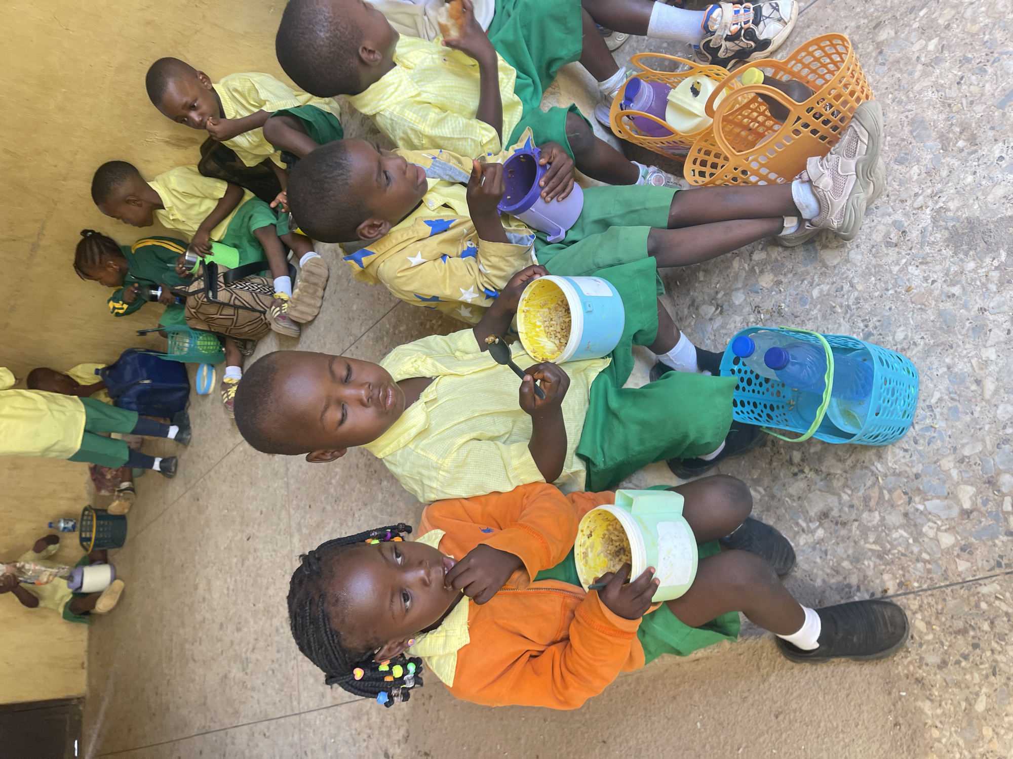 A group of children are sitting on a concrete surface, likely a schoolyard, in Akwanga, Nigeria. They are eating a meal out of plastic containers. Most of the children are wearing school uniforms that consist of yellow shirts and green shorts or skirts. Some of the children are using spoons to eat, while others are eating with their hands. Several plastic baskets and water bottles are on the ground next to the children. The lighting suggests the photo was taken during the day.