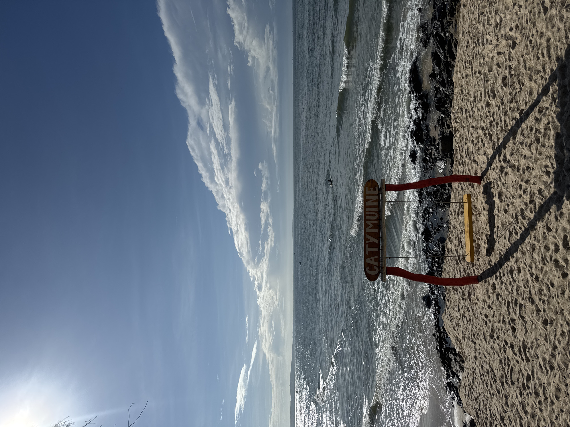 The image shows a wooden swing with a yellow seat positioned on a sandy beach. A red wooden frame supports the swing, and a sign above it reads "CATYMUINE". The beach is rocky in places, with waves breaking on the shore. The sea stretches out to the horizon, meeting a blue sky with scattered clouds. The sun is shining brightly, casting a shadow from the swing onto the sand. The scene appears to be in a coastal area, likely a beach resort, given the presence of the swing and signage. The specific location is the coast of Phan Thiết, Vietnam. There is one person visible in the water, seemingly swimming or surfing.