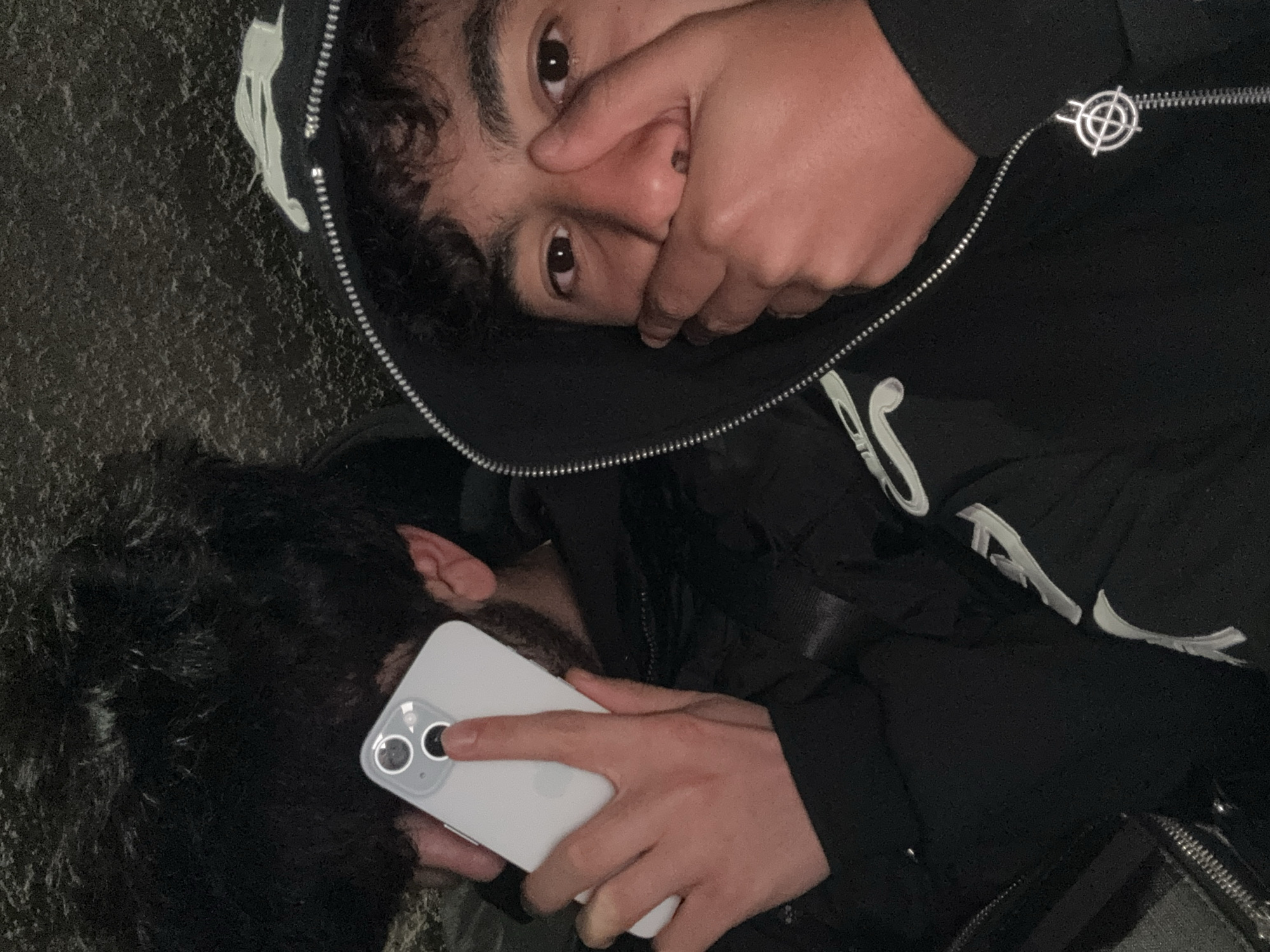 Two people are in the scene. One person, viewed from the shoulders up, is lying down with their face turned towards the camera, and their hand covering their mouth. They have dark curly hair, dark eyes, and are wearing a black hooded sweatshirt with white lettering. The zipper pull on the sweatshirt is a metal symbol resembling a target. The other person, viewed from the back of the head and shoulders, is holding a white smartphone with two camera lenses. The person has dark curly hair and a beard. They are wearing a black jacket or coat. The background is out of focus, but appears to be an urban environment. The location is Yerevan, Armenia.