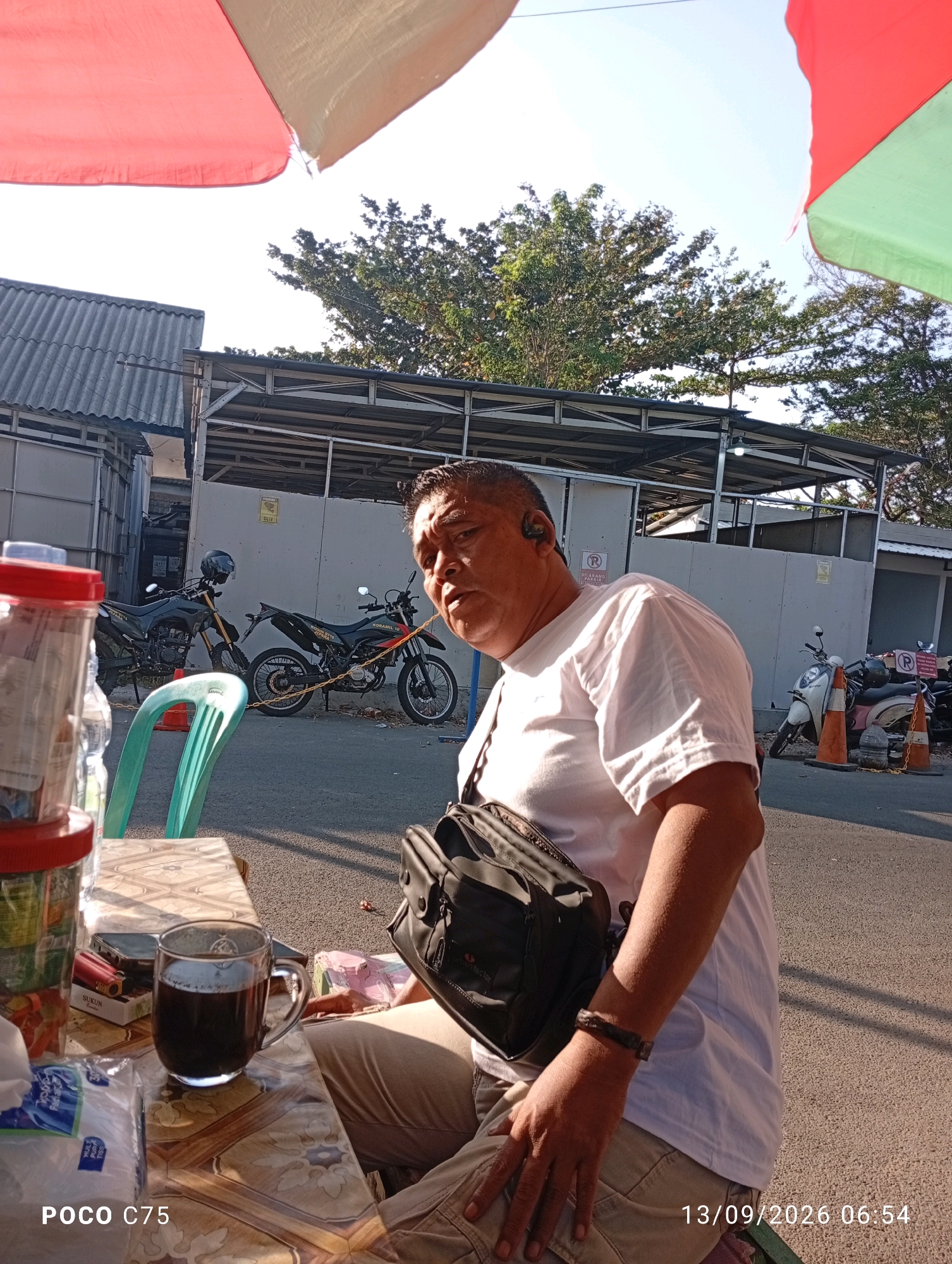A man wearing a white t-shirt and khaki pants is seated at a table outdoors, with a cup of dark liquid in front of him. He has a black crossbody bag slung over his shoulder and is wearing earbuds. In the background, two motorcycles are parked on a paved surface. A red and green umbrella provides shade. The scene appears to be on a street in Jepara, Indonesia, indicated by the timestamp in the lower right corner. The environment is urban, with buildings and trees visible.