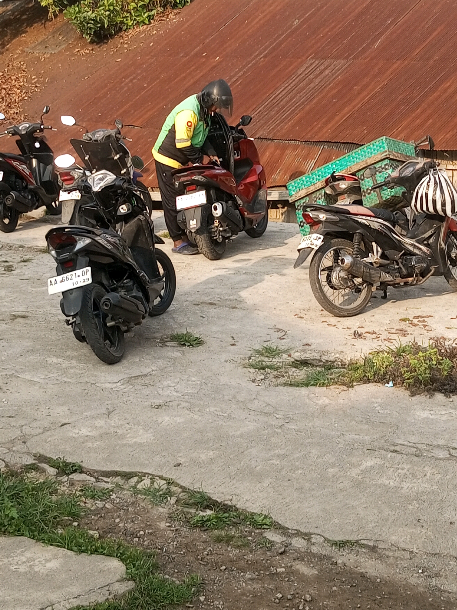 In Kalibeber, Indonesia, a person wearing a yellow and green jacket and black helmet is standing next to a red motorcycle, appearing to interact with it. Several other motorcycles are parked nearby, including a black one with a license plate reading "AA 6621 DP 10-29" and another black motorcycle with a striped bag attached to its handlebars. The scene is set on a concrete surface with patches of grass and dirt, and a weathered, rusty metal roof is visible in the background.