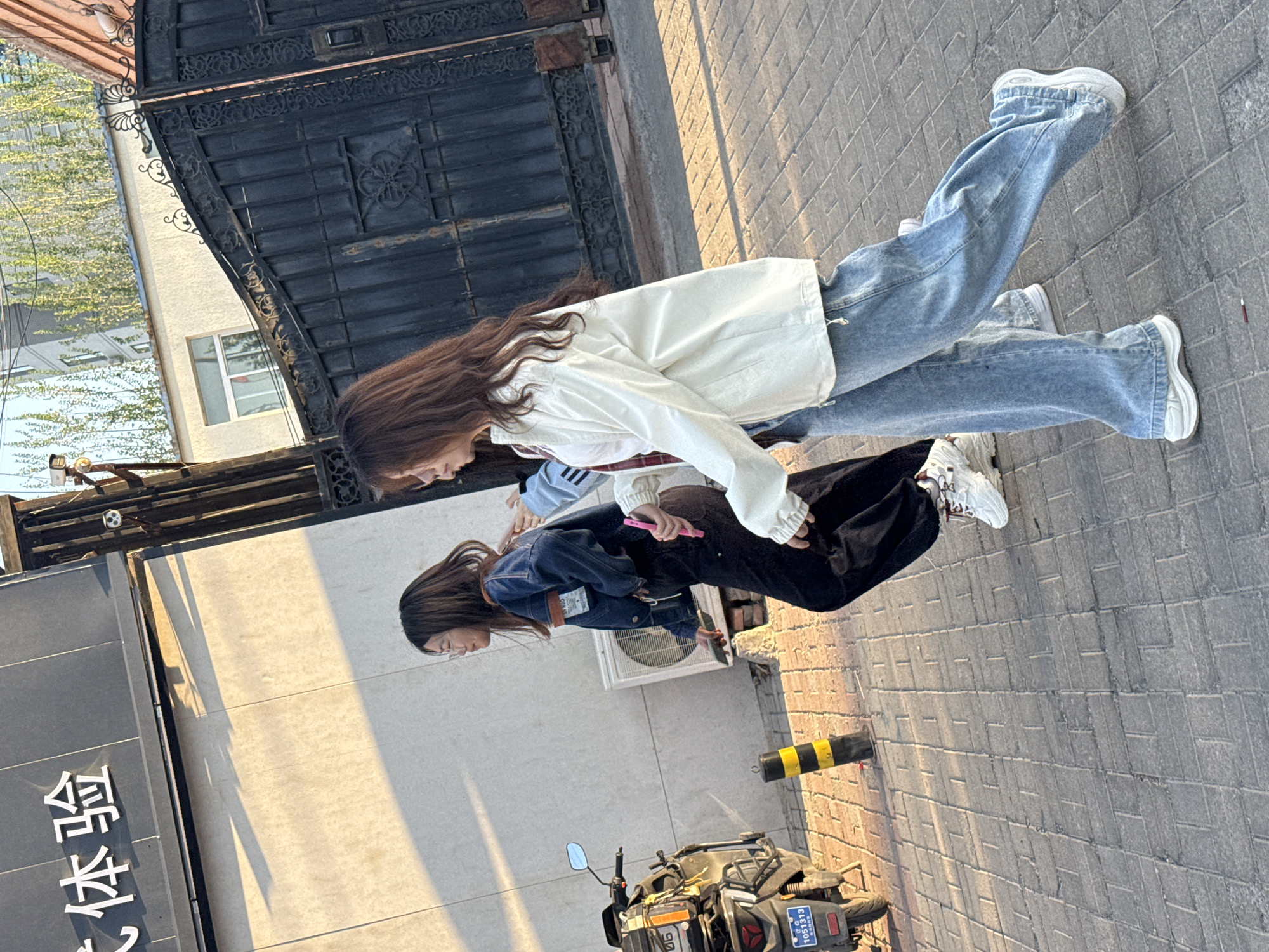 This image captures two young women on a paved street in Zhongtun, China, bathed in the warm, golden light of late afternoon or early evening, evident from the long shadows.
The woman in the foreground wears a white oversized jacket over a striped top, light blue wide-leg jeans, and white chunky sneakers. She holds a pink object, likely a smartphone, in her hand. Behind her, the second woman wears a dark blue denim jacket over a light-colored top with stripes on the sleeve, dark wide-leg pants, and white chunky sneakers. Both women have long brown hair and appear to be standing or walking casually, perhaps conversing.
The urban setting features a large, ornate black metal gate in the background, behind which a light-colored building with a window and some green foliage can be seen. To the far left, a dark sign with white Chinese characters reads "本色" (Běnsè), likely part of a business name such as "Bense Pub" (本色酒馆). On the right side of the street, a black and yellow striped bollard is visible next to a parked black motorcycle. The motorcycle features a blue Chinese license plate with the number "1051313." An outdoor air conditioning unit is mounted on a wall behind the second woman. The ground is paved with rectangular grey pavers. The scene depicts a candid moment on a sunny day in a Chinese urban environment.