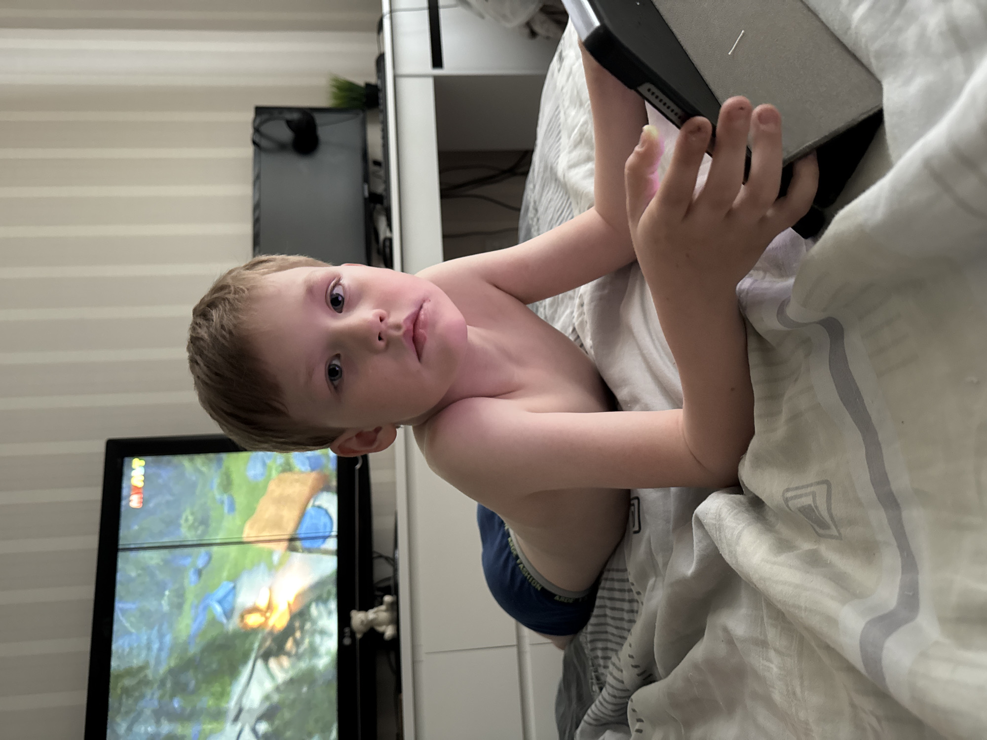 A young, light-skinned child is lying on a bed with light grey and white patterned sheets. The child is holding a tablet with a black and grey case. To the left, a television screen displays a brightly colored image, possibly a video game or animation. The child appears to be focused on the tablet. In the background, partially visible, is a white dresser and striped wallpaper. A small black object and some greenery sit on top of the dresser. The child's clothing is not fully visible, but a dark blue waistband is showing. Given the information provided and the image content, the location is likely Chaikovsky, Russia.