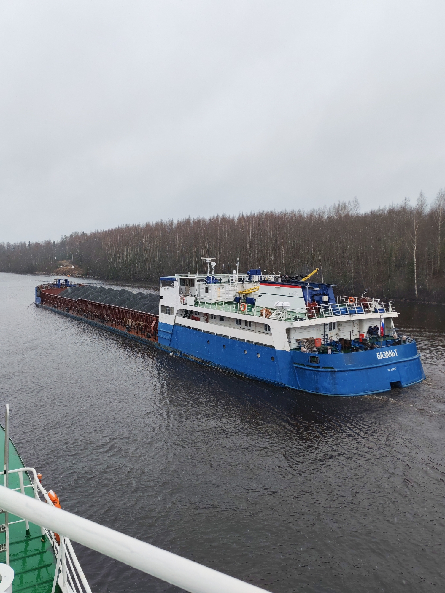 A barge named "BAZALT" is navigating a waterway in Volokov Most, Russia. The barge is blue and white and loaded with dark material, likely gravel or coal. Bare trees line the banks of the waterway under an overcast sky. A portion of another boat is visible in the foreground, suggesting the image was taken from a moving vessel.