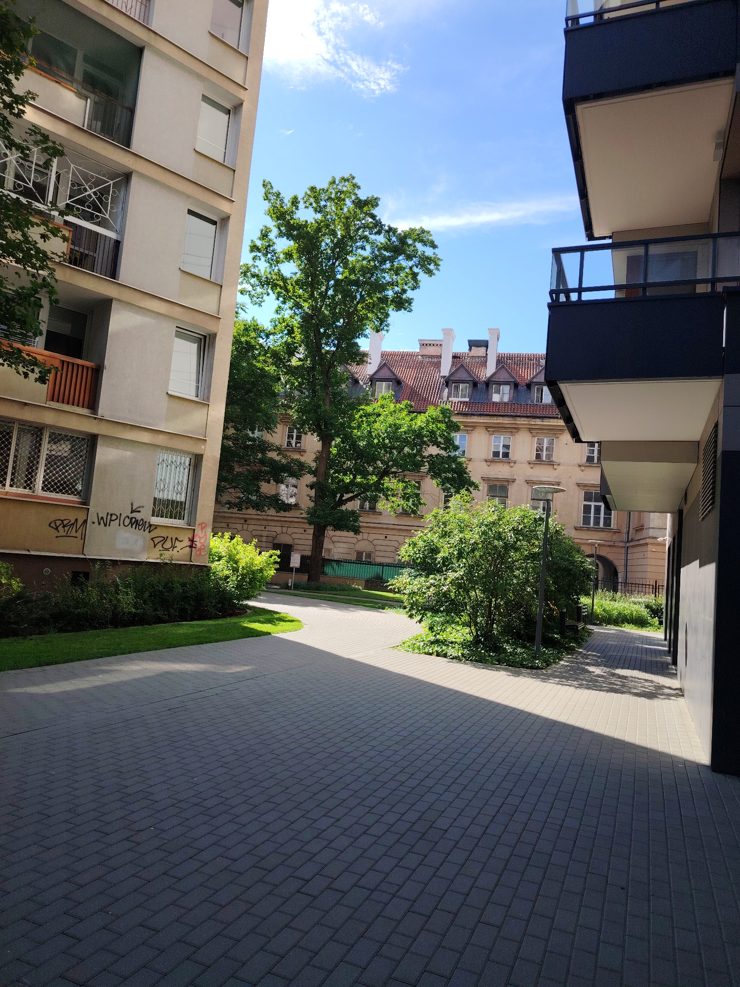 The image shows a courtyard area in front of residential buildings in Warsaw, Poland. On the left is a light-colored, multi-story building with balconies and windows, some of which have metal grates. The lower portion of this building has graffiti. To the right is a modern building with dark balconies. A paved walkway runs through the center of the image, bordered by grass and green bushes. In the background, an older, light-colored building with a red-tiled roof and multiple windows can be seen. Several mature trees with green foliage are present in the courtyard. The sky is blue with scattered clouds, indicating daytime. The scene appears to be a communal outdoor space within a residential complex.