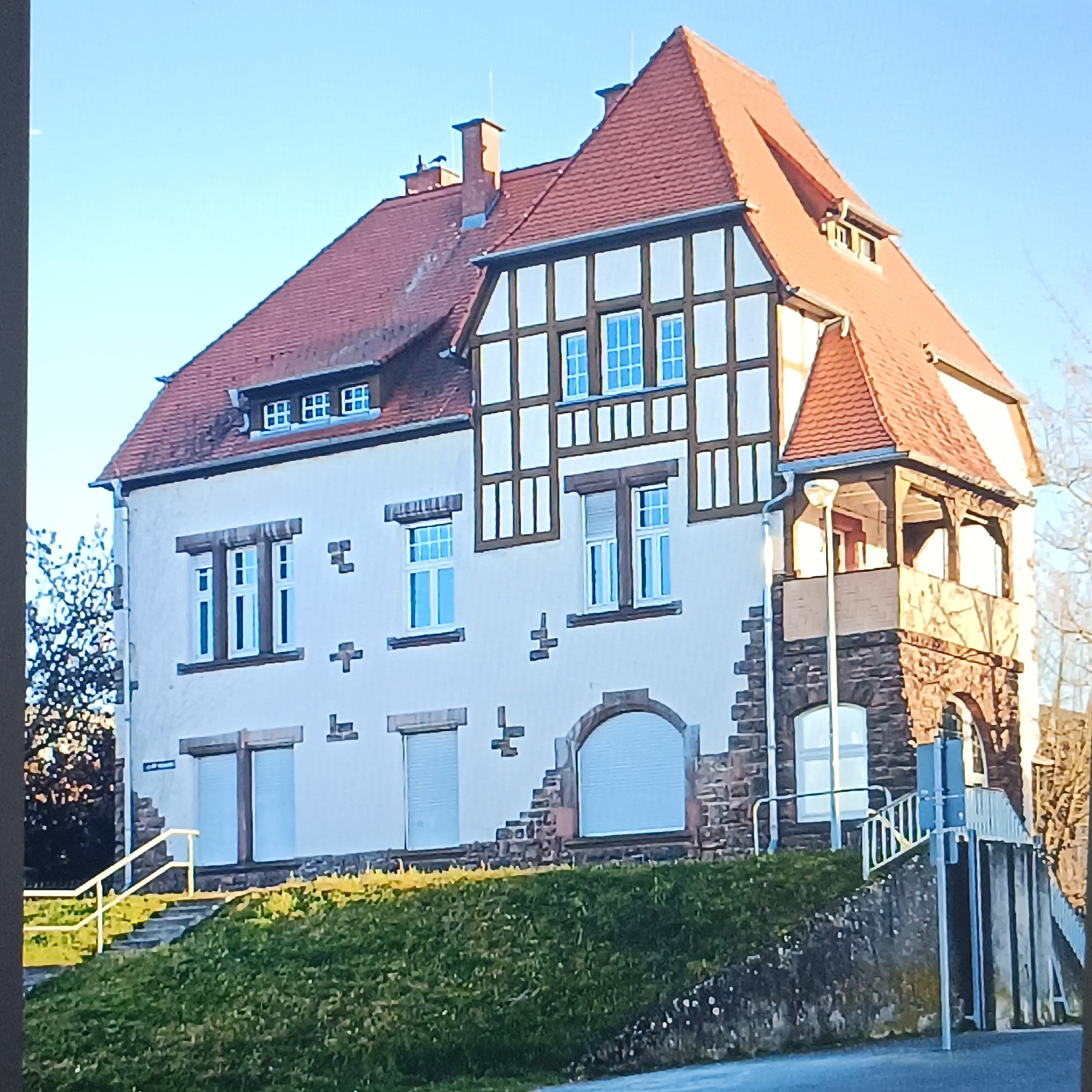 This image captures a building in Rüsselsheim am Main, Germany, under a clear blue sky. The building exhibits a distinctive architectural style with a red-tiled, multi-gabled roof. The facade is a combination of white stucco and timber framing with brown wooden beams creating a grid pattern. Several windows are visible, some with white frames and blue panes, and others with roll-down shutters. The lower portion of the building is constructed with reddish-brown stone. A small, covered balcony with wooden columns juts out from the right side of the structure. In front of the building, a grassy slope leads down to a paved area. A metal railing with stairs ascends the slope on the left side, and another set of stairs with railings is visible on the right side, partially obscured by vegetation. A street runs in the foreground, and a lamppost stands near the right-side stairs. No people are present in the image.