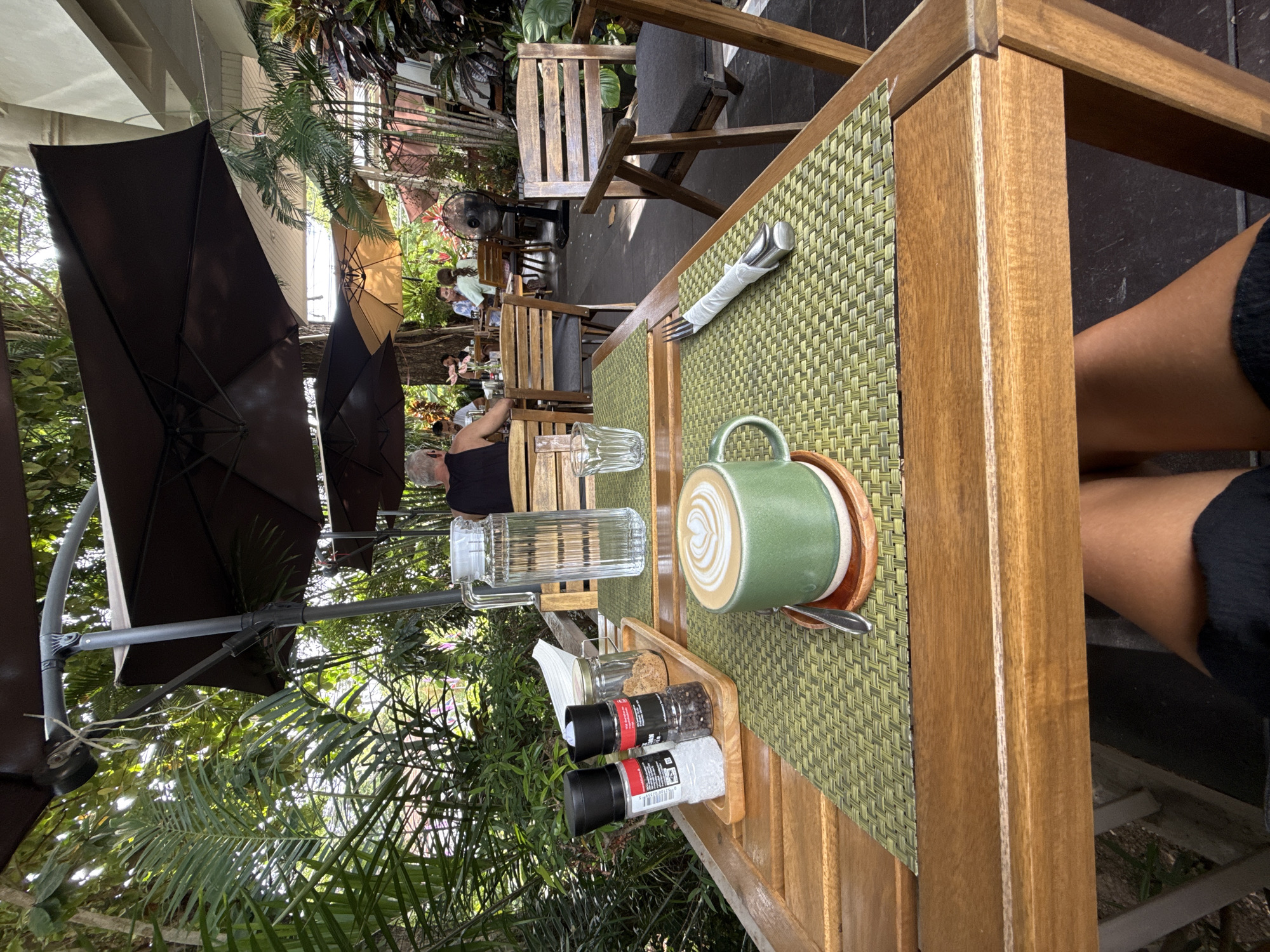 The image depicts an outdoor dining scene at a cafe or restaurant in Baan Chaweng Noi, Thailand. A wooden table in the foreground displays a green mug of latte art coffee, salt and pepper grinders, and a small wooden tray with sugar. A glass pitcher of water and a fork wrapped in a napkin are also on the table. In the background, several people are seated at other tables, enjoying their meals and drinks. The setting is lush with tropical foliage and covered by large umbrellas, suggesting a warm climate. The overall atmosphere appears relaxed and leisurely.