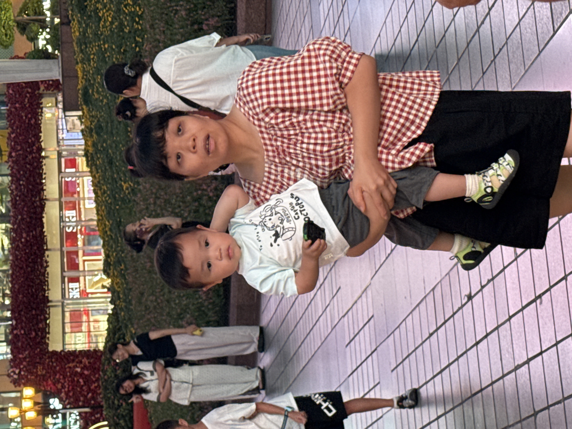 In Yuzhong District, China, a woman with dark hair and a red and white checkered shirt is holding a young child. The child, wearing a light blue t-shirt with a cartoon character and dark shorts, is looking towards the camera. The woman's gaze is also directed at the camera. They are standing on a patterned paved ground with other people in the background. To the left, a young boy in black shorts with white lettering and a white t-shirt is visible. The background features a building with large windows and a facade decorated with red floral arrangements and green plants. The lighting suggests it is daytime.