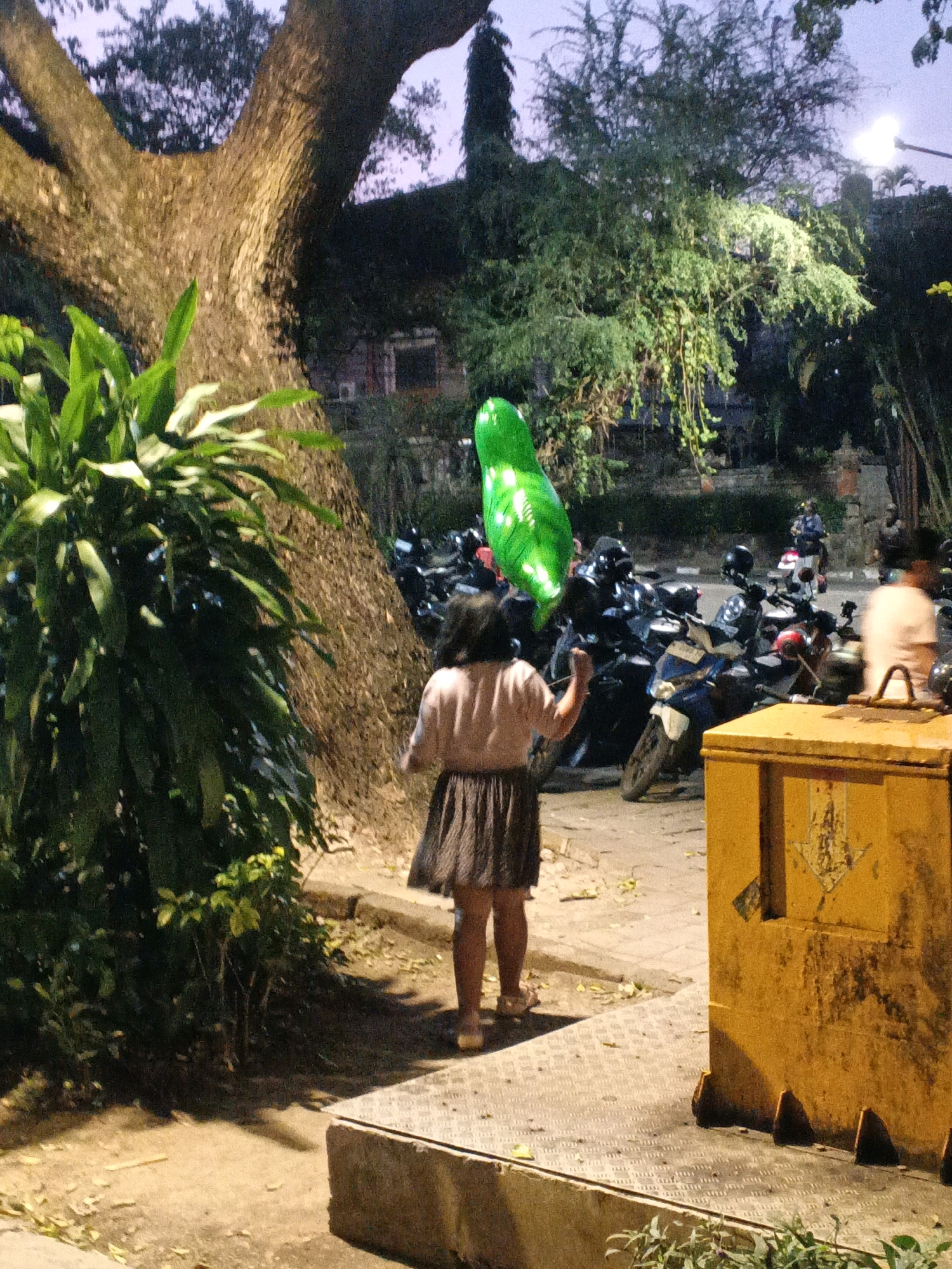 In Denpasar, Indonesia, a young girl with dark hair is walking away from the camera, holding a large, shiny green balloon. She is wearing a light pink short-sleeved shirt and a dark, pleated skirt, with light-colored sandals on her feet. The girl is on a paved area, next to a large, textured yellow trash bin. Behind her, a row of parked motorcycles are visible, some with helmets on them. A large tree with rough bark and dense green foliage is to her left, and other trees and vegetation are present in the background. The lighting suggests it is evening or dusk.