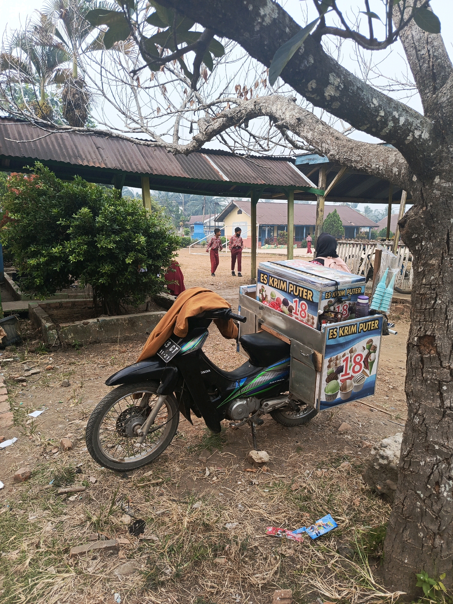 The image depicts an outdoor scene in a village or schoolyard in Megang Sakti II, Indonesia. A black motorcycle with an ice cream cart attached to its rear is parked on a dirt and grassy ground. The ice cream cart is decorated with images of ice cream cones and the text "ES KRIM PUTER" and the number "18". A brown jacket is draped over the motorcycle's seat. In the background, there are several buildings with corrugated metal roofs and what appear to be school children in uniform walking. The environment is somewhat unkempt, with dry grass, scattered leaves, and some debris on the ground. A large tree trunk is visible in the foreground on the right side of the image.