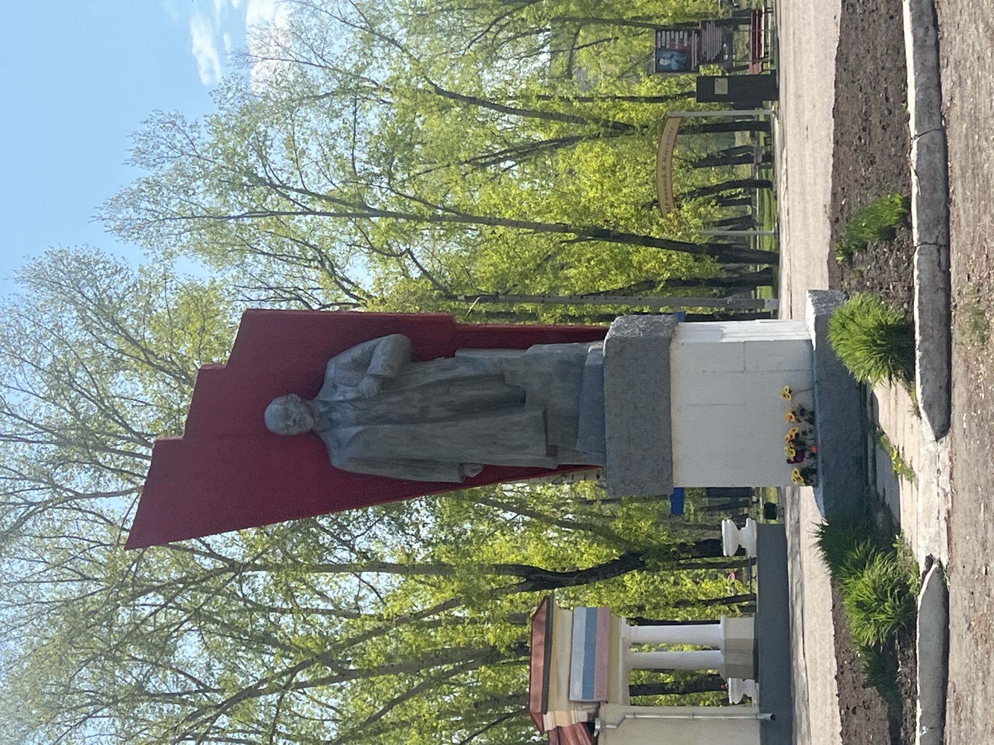 The image shows a statue of Vladimir Lenin in an outdoor setting, likely a park or square in an unidentified city. The statue is made of stone and features a red backdrop in the shape of a flag. The statue is positioned in front of green trees with new leaves. A white building with columns is visible below the statue. A paved walkway runs alongside the statue base with a strip of cultivated ground to its side. Yellow flowers have been placed at the base of the statue. In the background, there's a signpost and more trees. The sky is blue with few clouds.