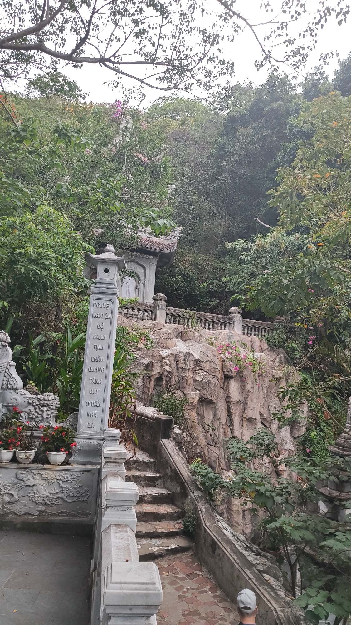The image shows a stone staircase leading up a rocky incline, surrounded by lush green foliage. A traditional-style pavilion with an ornate roof sits atop the rocky formation. In the foreground, a stone lantern-style monument stands next to a wall with carved decorative elements and potted flowers. The scene appears to be a temple or historical site. The text on the stone monument is in Vietnamese. The overall atmosphere is serene and naturalistic, suggesting a location in a mountainous or hilly area within a city. Based on the style of architecture and the presence of Vietnamese text, this is likely a location within Vietnam, possibly a temple complex or cultural site in or near the city of Đà Nẵng, Vietnam. The image captures a daytime scene, with overcast sky visible through the trees.