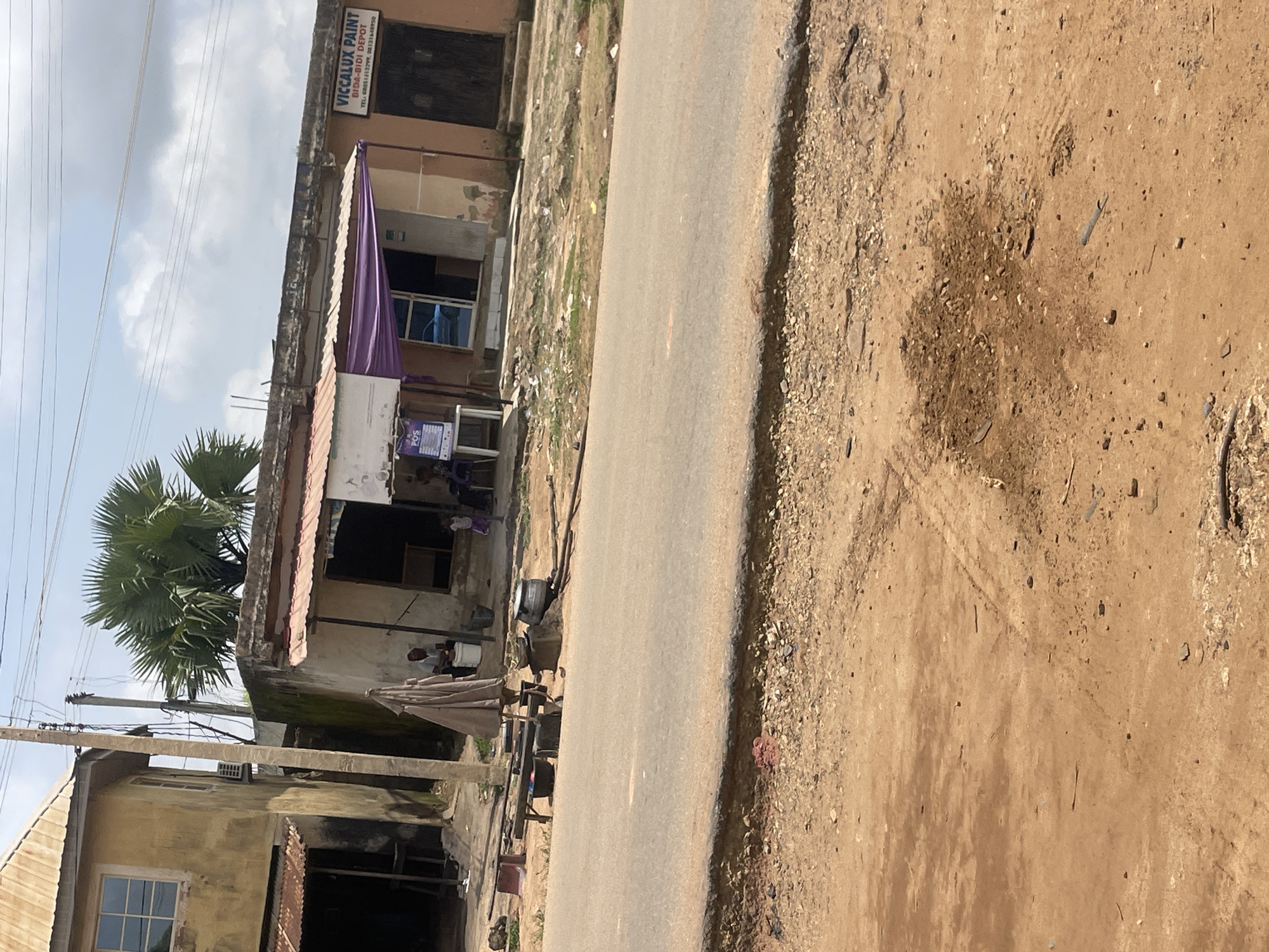 The image shows a street scene in Unguwar Saya, Nigeria. A two-story building with a weathered yellow facade and a corrugated metal roof is visible on the left, with a window on the upper floor and a covered entrance on the ground level. Adjacent to it, a single-story building has a similar corrugated metal roof. This building features a purple awning over an open storefront. To the right of the storefront, a sign reads "VICCALUX PAINT BYDA-BIDI DEPOT". There are dark wooden doors on the far right of this building.
In front of the buildings, a paved road runs horizontally across the image. On the side of the road, near the buildings, several objects are present. A person is seated on a low stool next to a white bucket. Nearby, there are cooking pots, a table with some items on it, and what appears to be laundry. A large umbrella is positioned over the table. Further to the right, two children are visible, one standing and the other seated.
The ground in front of the road is composed of reddish-brown earth with scattered stones and some vegetation. In the foreground, tire tracks are imprinted in the dirt. The sky above is partly cloudy. The overall scene depicts a typical roadside environment with commercial activity and residential elements.