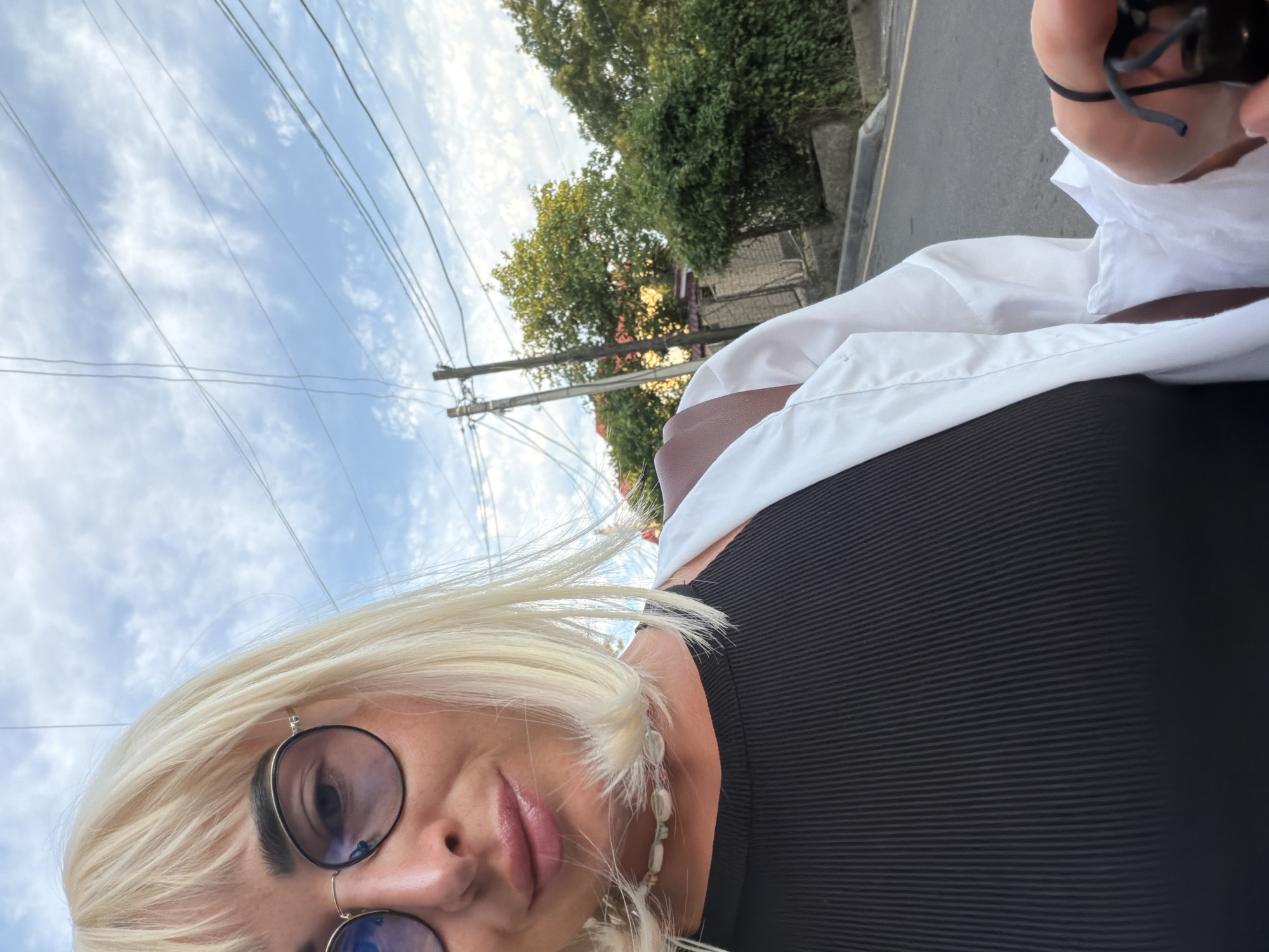 A selfie is taken outdoors in Sochi, Russia, with a cloudy sky and some power lines visible in the background. The subject, a woman with blonde hair and bangs, wears round glasses and a black ribbed top with a white open shirt over it. She also sports a beaded necklace. Her left hand holds a black face mask. The background shows a residential area with trees and a street. The lighting suggests it is either early morning or late afternoon.