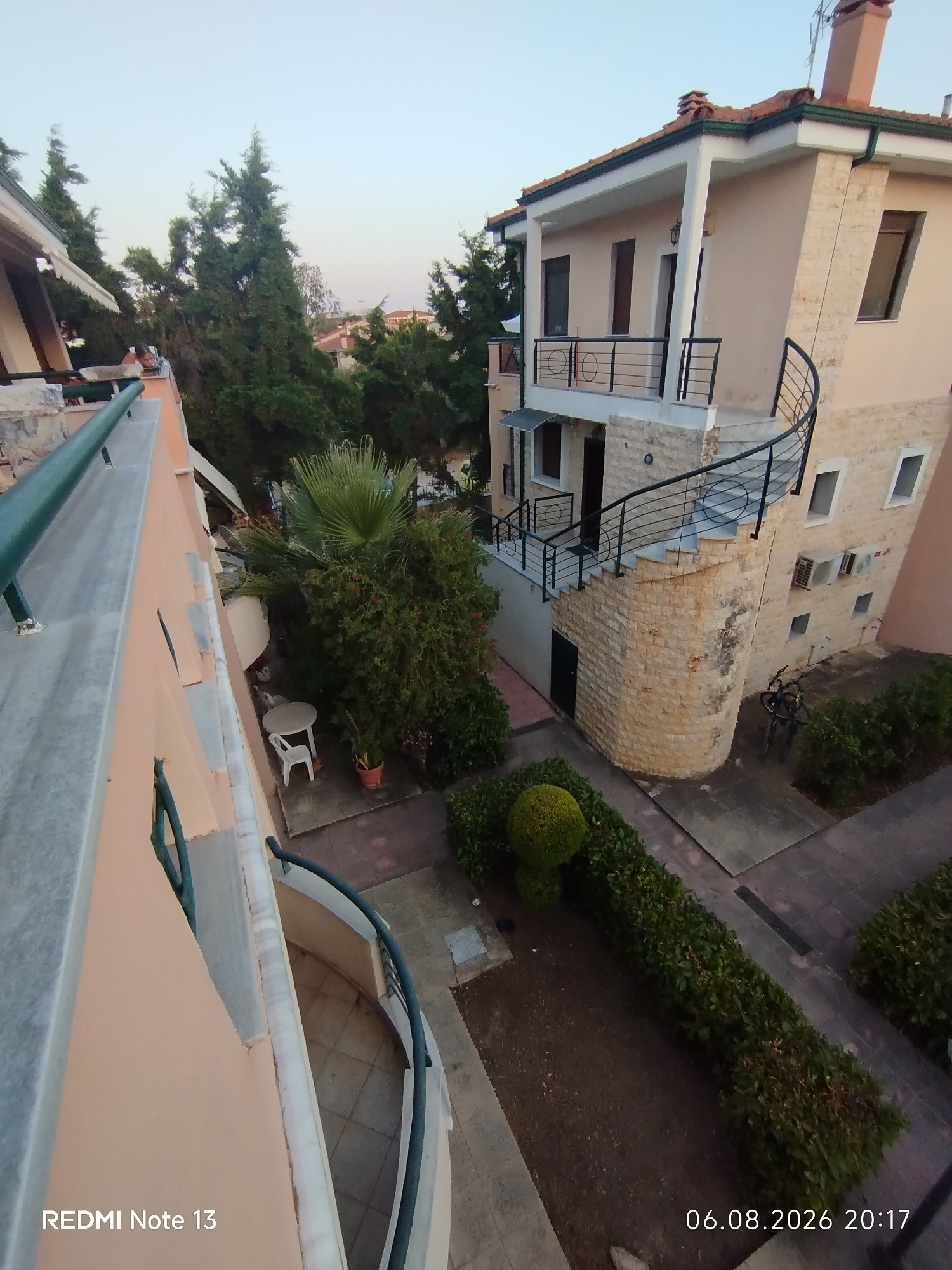 This is an outdoor, high-angle, medium shot of a multi-story building in Sithonia Municipal Unit, Greece. The building is constructed with a light-colored stone base and a peach-colored upper story. A winding external staircase with black metal railings leads up to the second floor. A small balcony is visible on the second floor, also with black metal railings. Two air conditioning units are mounted on the exterior wall. Two bicycles are parked in front of the building.
The surrounding area features green trees and manicured hedges. A small patio area with a round table and a white plastic chair is visible. The sky is clear and light blue, indicating it is either early morning or late afternoon. The image has a date stamp in the bottom right corner that reads "06.08.2026 20:17". The bottom left corner has a phone model indicator that reads "REDMI Note 13".