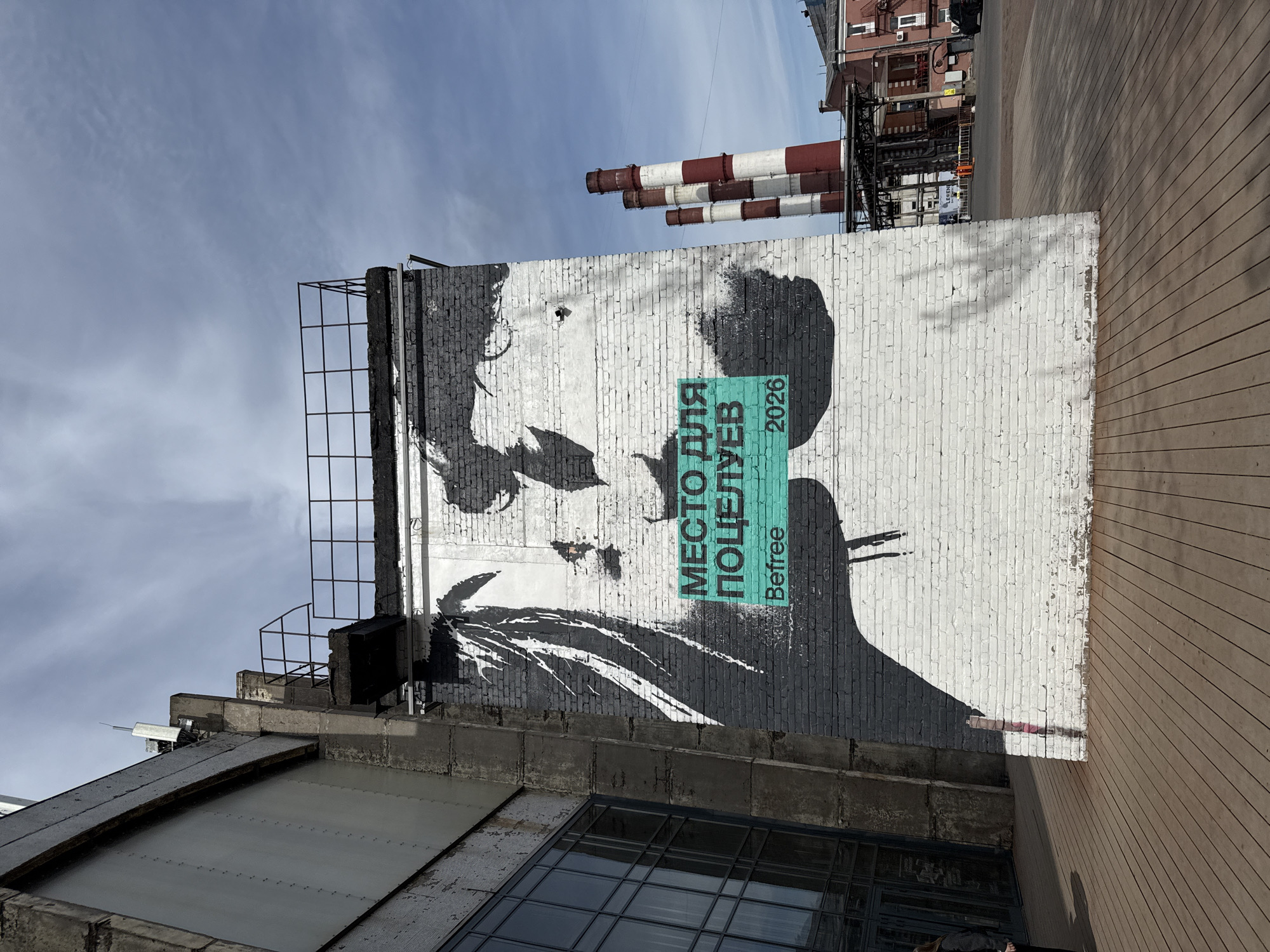 The image displays a large black and white mural of a stylized human face on a brick wall, situated in an urban or industrial outdoor setting. The photograph is oriented vertically, with a bright, partly cloudy sky dominating the left side, and a wooden-planked surface on the right.
The mural itself is a striking piece of street art, featuring a minimalist depiction of a face, possibly female, rendered with black brushstrokes and negative space on the white-painted brick. The brick surface shows some wear and texture, which integrates into the art's aesthetic. Overlaid on the mural, a prominent teal rectangular box contains Russian text. It reads "МЕСТО ДЛЯ ПОЦЕЛУЕВ" (MESTO DLYA POTSELUYEV), which translates to "PLACE FOR KISSES." Below this, the year "2026" is visible, along with the brand name "Befree."
In the background, positioned above the mural and behind other structures (relative to the vertical orientation), are several tall industrial chimneys with distinctive red and white stripes, characteristic of industrial facilities. Other buildings with brick facades are visible further in the background. A metal scaffolding or framework is attached to the upper left edge of the wall where the mural is painted, and parts of an adjacent building's roof with metal framing are also seen. The wooden surface on the right appears to be a deck or walkway, adding a contrasting element to the industrial backdrop.
The scene is well-lit by natural daylight, suggesting a clear day. No people or overt activities are present, giving the environment a serene and observational quality. The mural itself conveys a sense of contemplation or mystery, enhanced by its gritty urban setting.