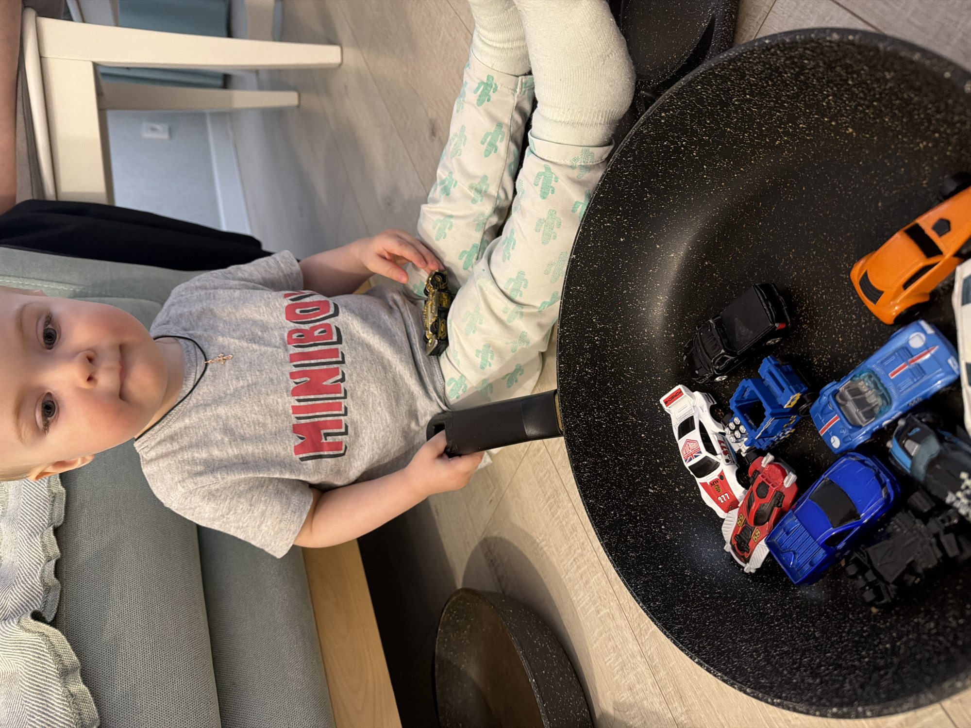 A young child sits on a light blue sofa wearing a gray t-shirt with "MINIBOY" printed on it, light green cactus-patterned pants, and white socks. The child holds a small, gold-colored toy car in their right hand and the handle of a large black frying pan in their left hand. The frying pan contains several toy cars of various colors (orange, black, white, red, and blue). The room has light-colored wooden flooring. The photograph's location is unknown and is not explicitly shown as being in Votkinsk, Russia.