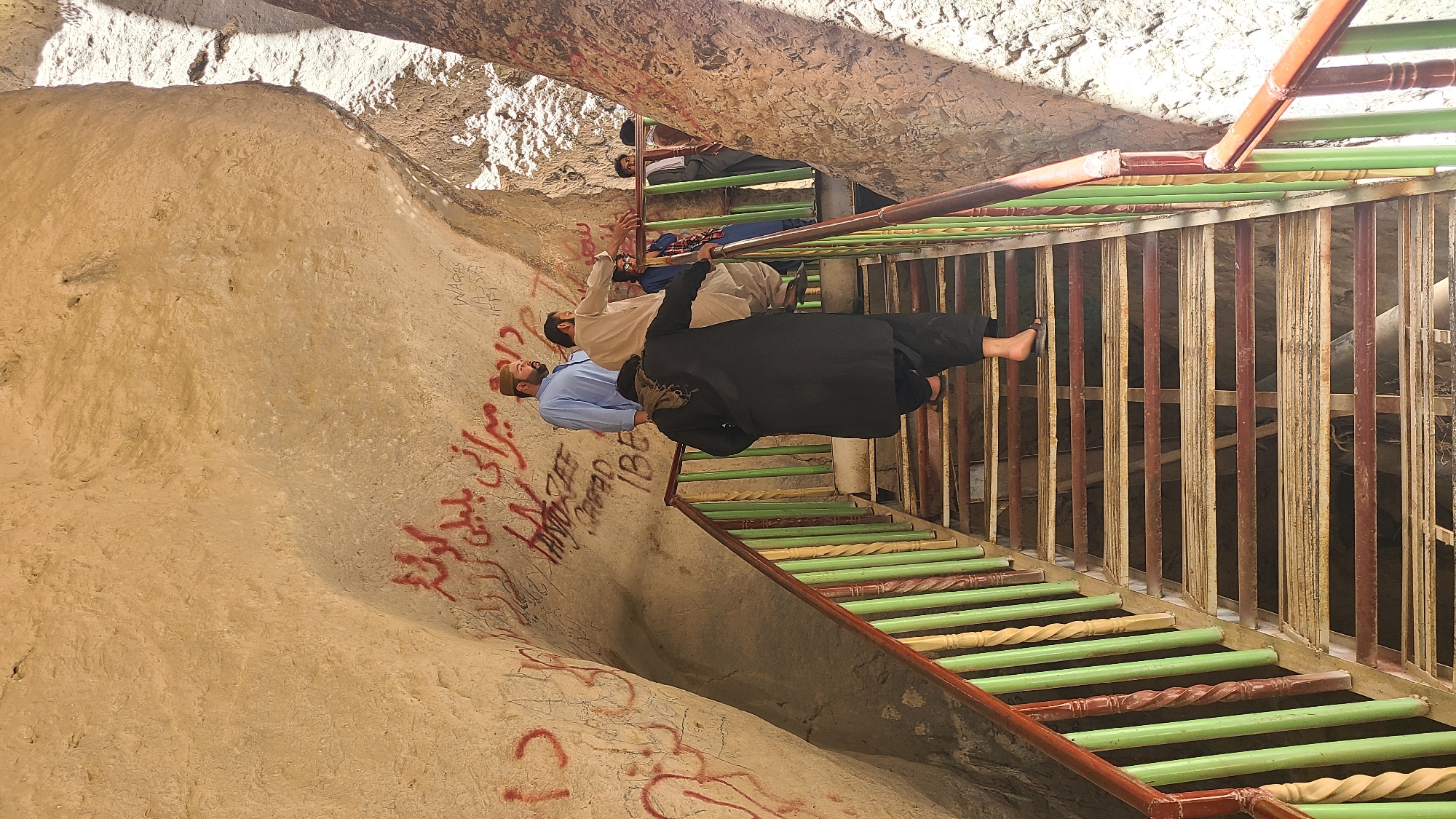Three men are on a stairway. The wall to the side is off-white and has some graffiti. The stairs are painted with light green and brown. The person closest to the viewer is wearing a black dress and has bare feet. There is metal fencing along the side of the stairs.