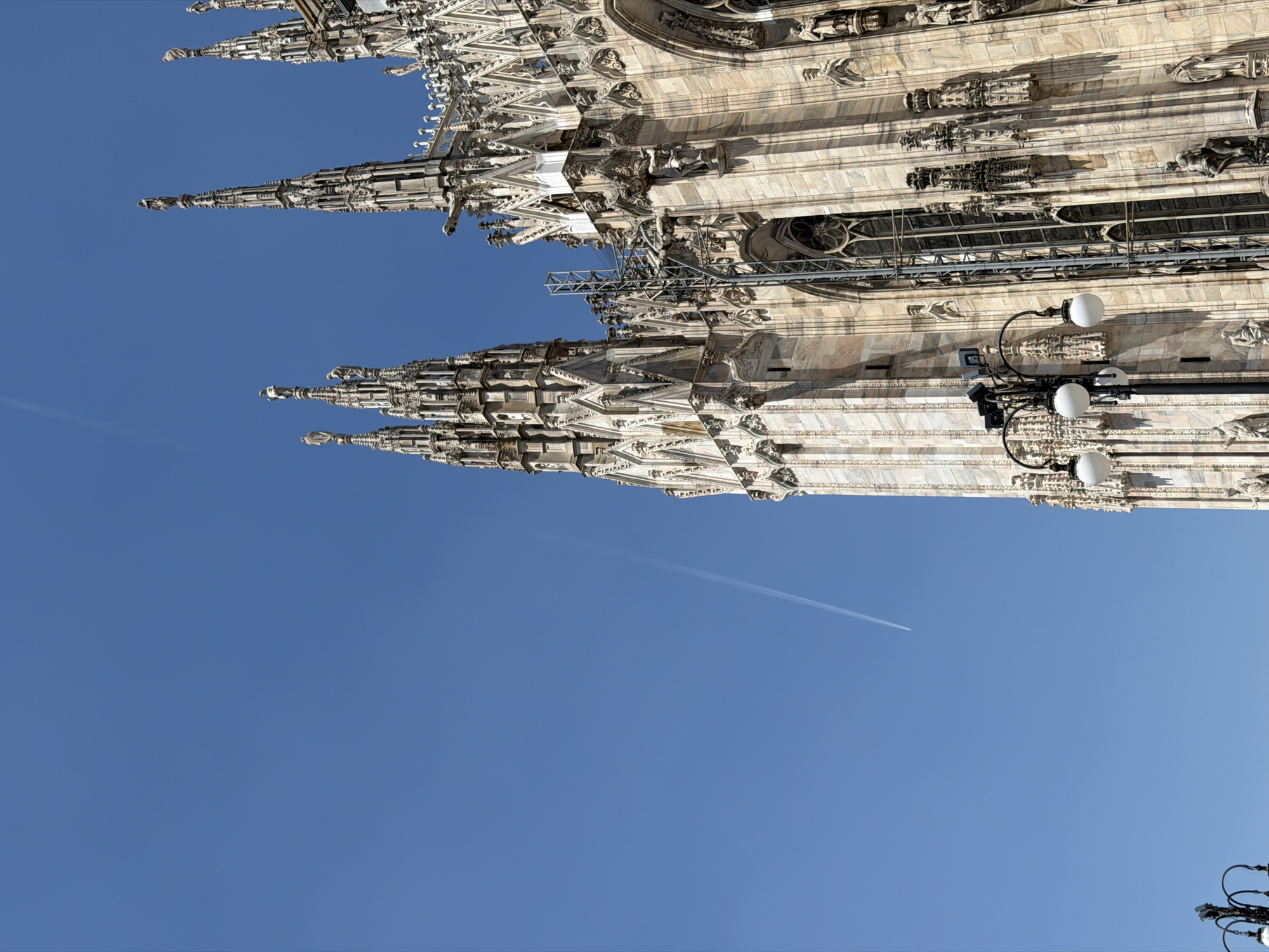 The image shows a view of the Duomo di Milano (Milan Cathedral) in Milan, Italy, from a low angle. The lower portion of the image is dominated by a clear, blue sky with a faint contrail visible. Above the sky, the elaborate spires and architectural details of the Duomo are prominently displayed. The facade is made of white marble, showing intricate carvings and sculptures. Some scaffolding is present on the structure. Street lights are attached to the side of the structure.