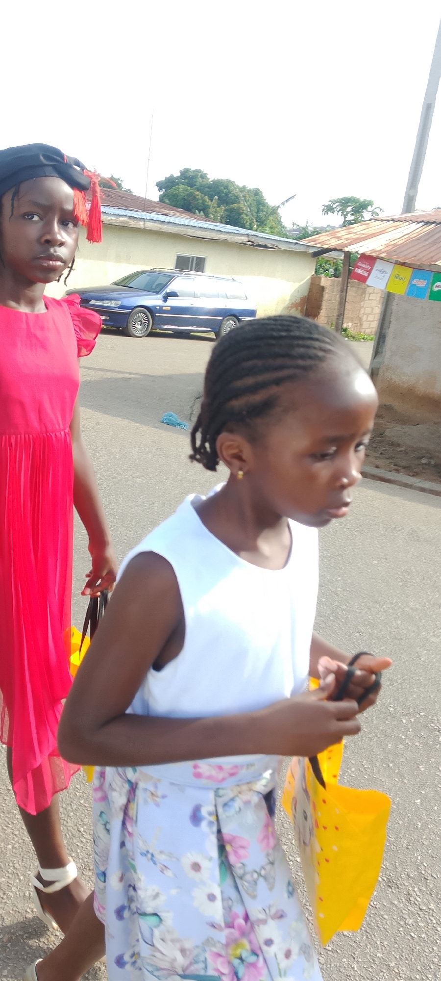 Two young girls are walking on a paved street. The girl in the foreground, on the right, has her hair braided in cornrows and is wearing a white sleeveless top and a light blue dress with a floral pattern. She is carrying a yellow bag in her right hand. She is looking to her right. The girl in the background, on the left, is wearing a bright red dress and a black cap with red tassels. Her hair is braided and she appears to be looking to her left. A blue station wagon is parked on the side of the street behind the girls. Buildings with corrugated metal roofs are visible in the background. The sky is bright and clear, suggesting it is daytime.