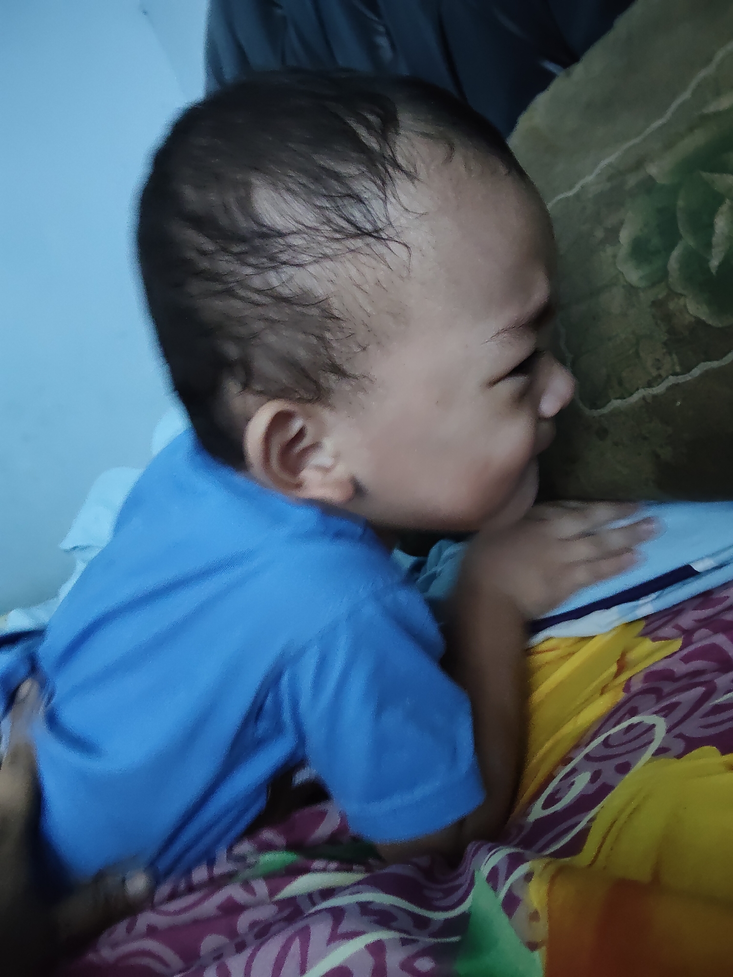 In Tanah Grogot, Indonesia, a close-up, side-profile view captures a young child lying down, facing to the right, with their head resting on a pillow or cushion. The child is wearing a bright blue t-shirt. Their skin appears light-toned, and their hair is dark and slightly damp. The child's left arm is visible, with their hand resting on a folded blue and white striped fabric, likely part of bedding. The background includes a dark, indistinct object at the top, possibly clothing or furniture, and a patterned blanket or sheet beneath the child, featuring swirling designs in shades of purple, green, and yellow. A hand is visible in the lower left of the frame, suggesting someone is holding or supporting the child. The lighting is moderate, and the overall image has a slightly soft focus.