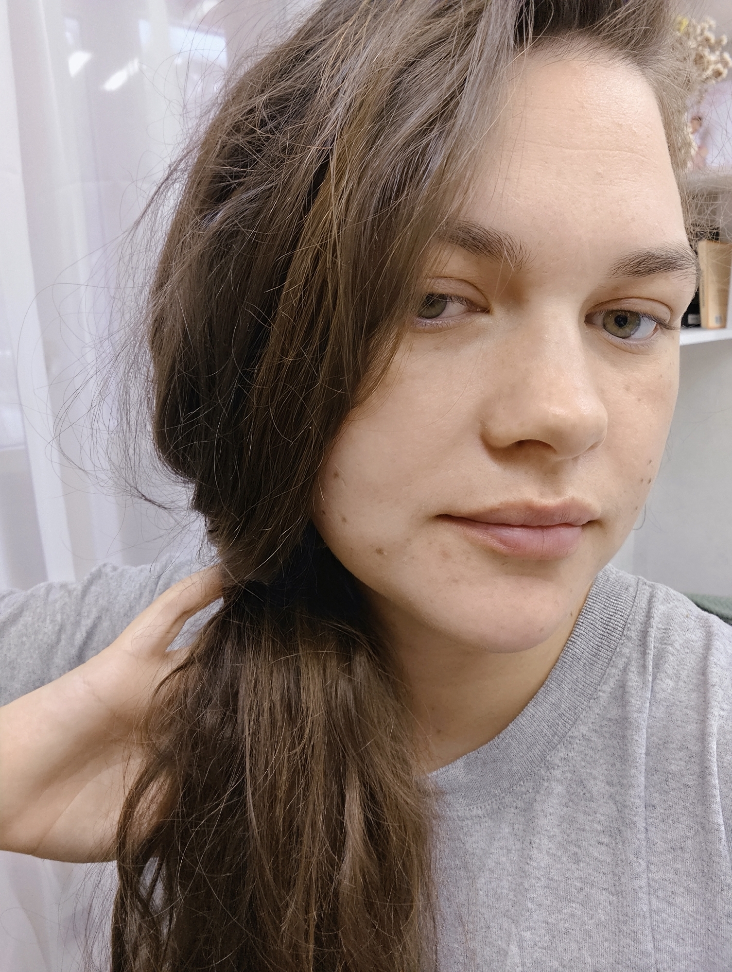 A close-up, front-facing portrait of a young adult female is depicted in an indoor setting in Chaikovsky, Russia. The subject has fair skin exhibiting minor blemishes and visible pores, green-hazel eyes with natural dark lashes, and medium-brown hair that appears disheveled, falling loosely over her shoulders. Her lips are closed, presenting a neutral expression. She is wearing a grey crew-neck t-shirt. Her right hand is visible, with fingers lightly grasping a portion of her hair on the left side of the frame. The background consists of a light-colored wall or partition, with a sheer white curtain visible on the left and a shelf displaying indistinct objects, including dried foliage, on the right.