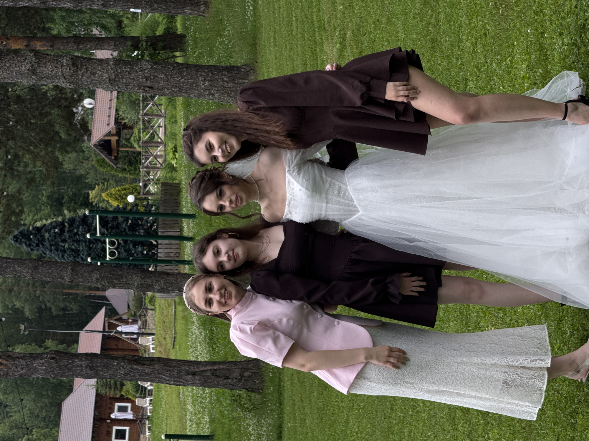 Four young women are posing for a photograph on a grassy lawn. The woman on the far left is wearing a light pink jacket over a white lace skirt and silver sandals. The woman next to her is wearing a dark brown dress and black heels. The woman in the center is wearing a white wedding dress with a veil. The woman on the far right is wearing a dark brown dress and black heels. They are standing in front of trees and some wooden buildings, which appear to be part of a resort or park. The overall environment is green and lush, suggesting a summer or late spring day. The women appear to be happy and are smiling for the camera. This scene is likely taking place at the "Novyy Svet" resort in Votkinsk, Russia, given the style of the wooden buildings and the natural landscape.