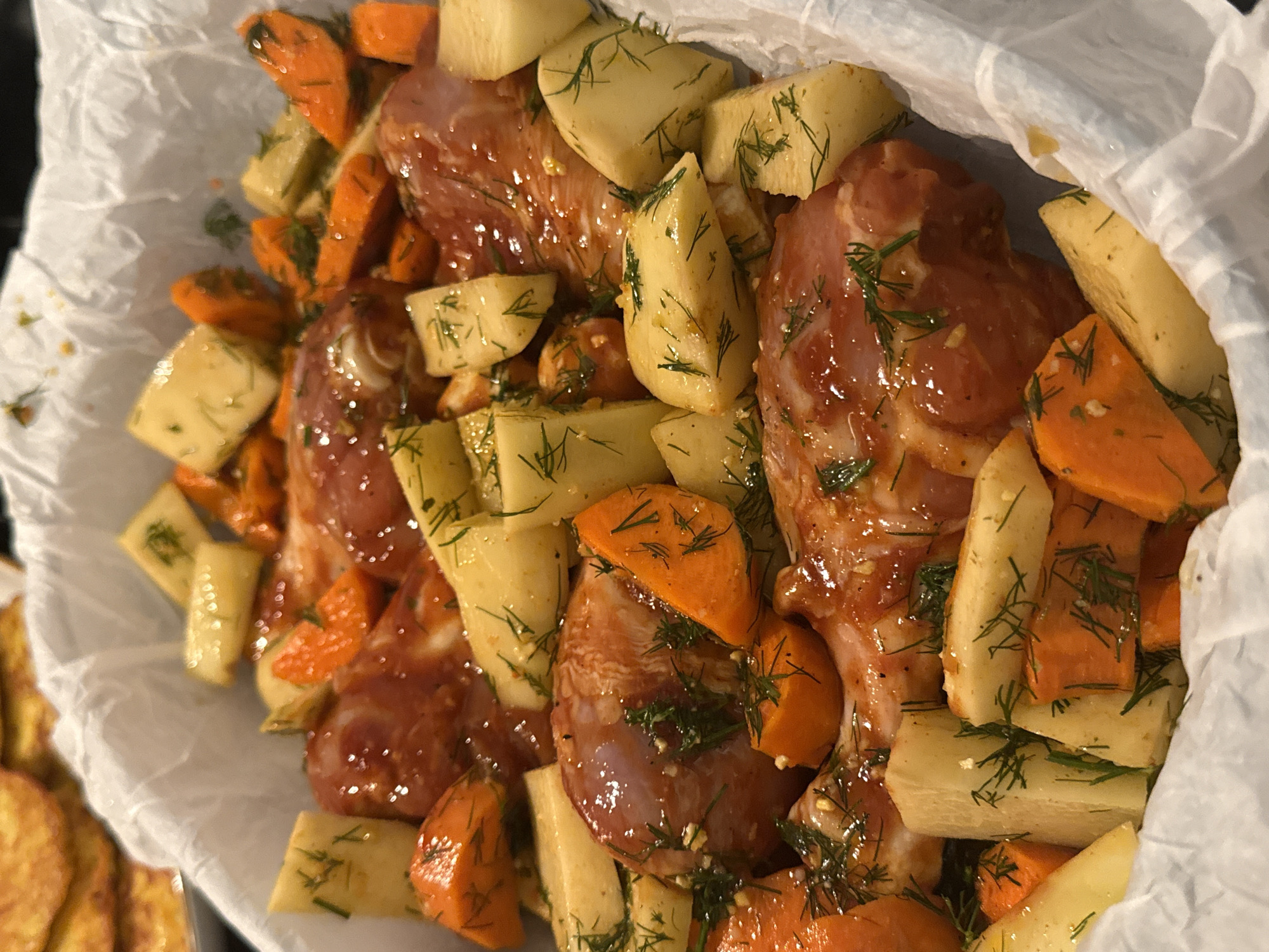 The image shows a close-up view of a roasting pan lined with white parchment paper. Inside the pan are pieces of raw chicken coated in a reddish-brown sauce, interspersed with chunks of potatoes and carrots. Fresh dill is sprinkled over the ingredients. The chicken pieces appear to be thighs or drumsticks, and the vegetables are cut into bite-sized pieces. The overall presentation suggests a meal being prepared for cooking. There is no information in the image to suggest a specific location in New York City, United States of America.