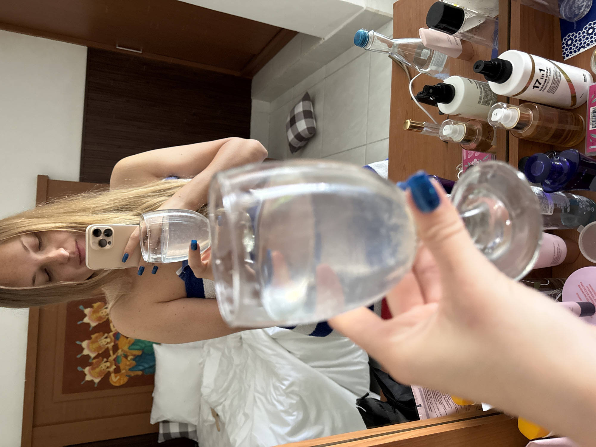 A blonde woman is taking a mirror selfie in a bedroom. She is holding a glass of water and her phone in her hands. She is wearing a blue and white striped towel. The room has a bed, a wooden wardrobe, and a dresser with various toiletries. A framed artwork depicting three figures is visible on the wall behind the bed. A small checkered pillow is on the floor near the wardrobe. The dresser top is cluttered with bottles of perfume, skincare products, and a spray bottle labeled "17 in 1 CREAM SPRAY PERFECT HAIR". The image appears to have been taken in Denpasar, Indonesia, as indicated by the presence of local artwork and possibly the style of the hotel room.