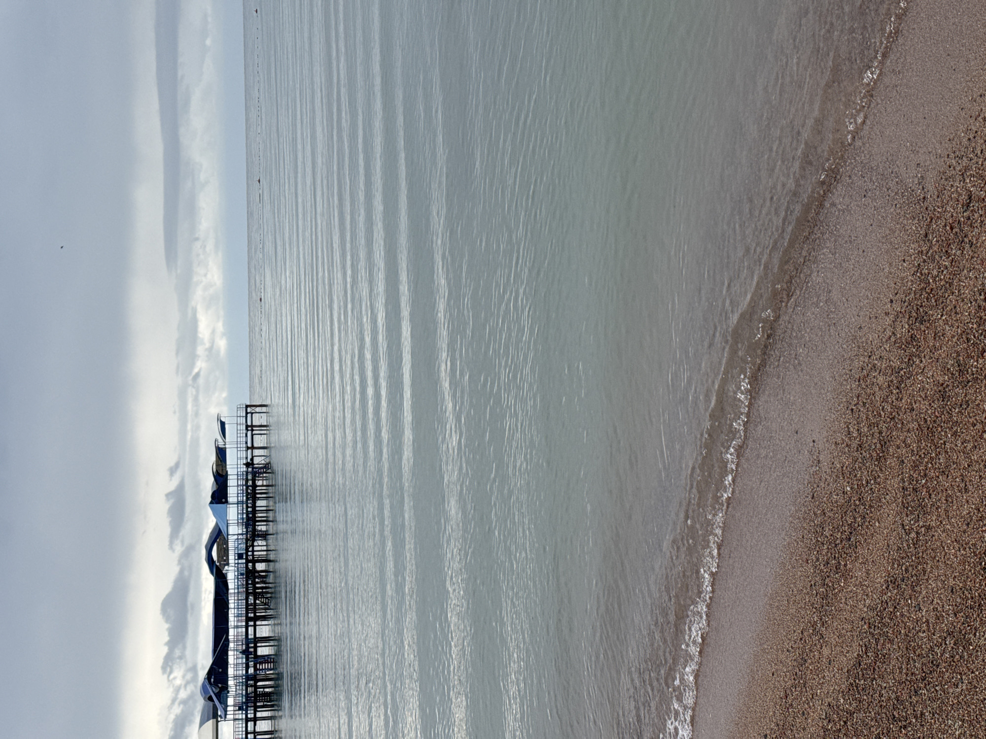A view of a beach and a pier over the Issyk-Kul Lake in Sary-Oy, Kyrgyzstan. The pier, constructed of wood with blue roofing, extends into the calm, rippling water. The beach is covered in small, brown pebbles. The sky is overcast with a muted, grey tone. The image is a wide shot capturing the expanse of the lake and the serene atmosphere.