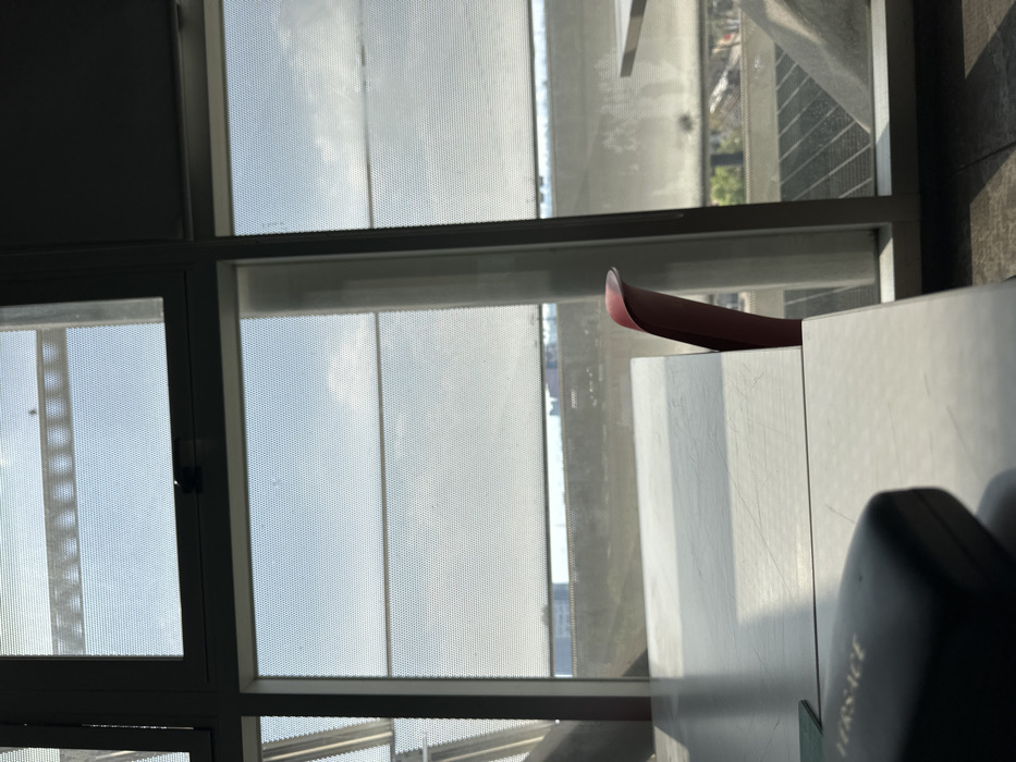 A partial view through a window with a dotted screen shows a cityscape. A maroon shoe rests atop a white surface with vertical scratches. The bottom right reveals a dark case with the word "VERSACE" inscribed on its side. The setting appears indoor, likely in a building with a view.