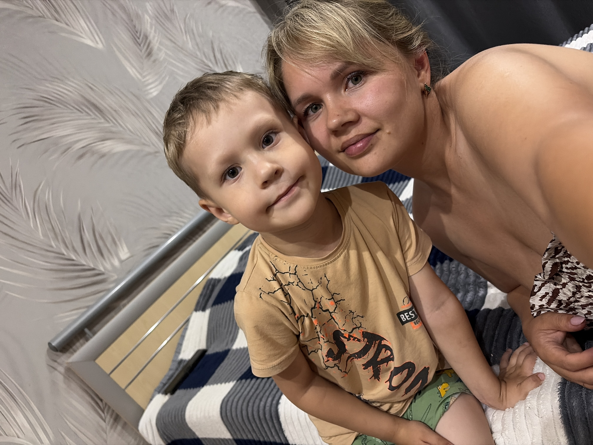 In this medium shot selfie, a young boy and an adult woman, likely his mother, are close together, both looking towards the camera with soft smiles. The boy, with short blonde hair and large expressive eyes, wears a tan t-shirt with a graphic design of cracked lines and what appears to be black text. He is also wearing green shorts with a dinosaur pattern. The woman, also with blonde hair tied back and bangs framing her face, has a warm expression and is wearing a small green earring. She is bare-shouldered, suggesting a casual or home setting.
They are positioned on a bed with a black, white, and grey striped comforter. Behind them, the wall is covered in a textured wallpaper with a light-colored, repeating feather or leaf pattern. A metal railing, possibly from a bed frame or crib, runs along the left side of the image. The lighting suggests it is daytime, likely indoors, given the intimate setting and absence of natural light cues. The overall mood is one of tenderness and connection between the subjects. No visible text other than on the boy's t-shirt, which is partially obscured. The location context of Chaikovsky, Russia, implies a domestic scene in a residential setting.