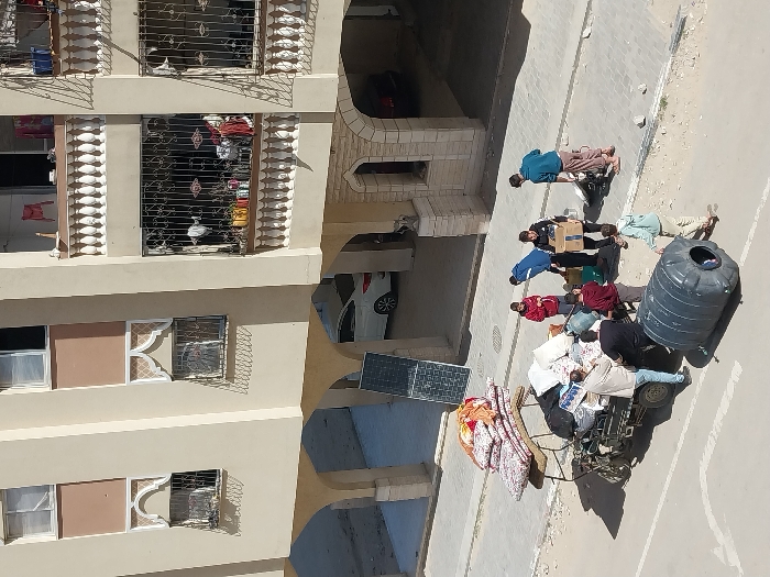 The image captures a street-level scene in Khan Yunis, Palestinian Territory, in front of a multi-story residential building with light beige facades and brown architectural panels. The building features multiple windows, some with ornate arched frames and decorative iron grilles, and balconies with white balustrades and black iron railings. On the paved sidewalk adjacent to the building, a large, folded black solar panel rests vertically against the wall.
On the asphalt road in the foreground, a three-wheeled motorized vehicle with a flatbed is heavily laden with various household items. These include a stack of approximately seven floral-patterned mattresses, numerous large bundles of white and colored textiles, and several bags. Eight individuals, primarily male adults and young adults, are positioned around the vehicle and on the adjacent paved area. Actions involve several men handling cardboard boxes and interacting with the loaded goods on the vehicle. One man in a light shirt is near the left side of the vehicle, while another in a black shirt stands beside him. Closer to the right, a man in a blue jacket holds a cardboard box, and another individual in a red shirt stands nearby. A large, black, cylindrical plastic water tank lies horizontally on the asphalt road in the immediate foreground, to the right of the vehicle. A younger individual in a light hoodie walks towards the right, away from the main group. A white car is visible parked under an arched recess of the building's ground floor. Shadows are sharp, indicating direct sunlight.