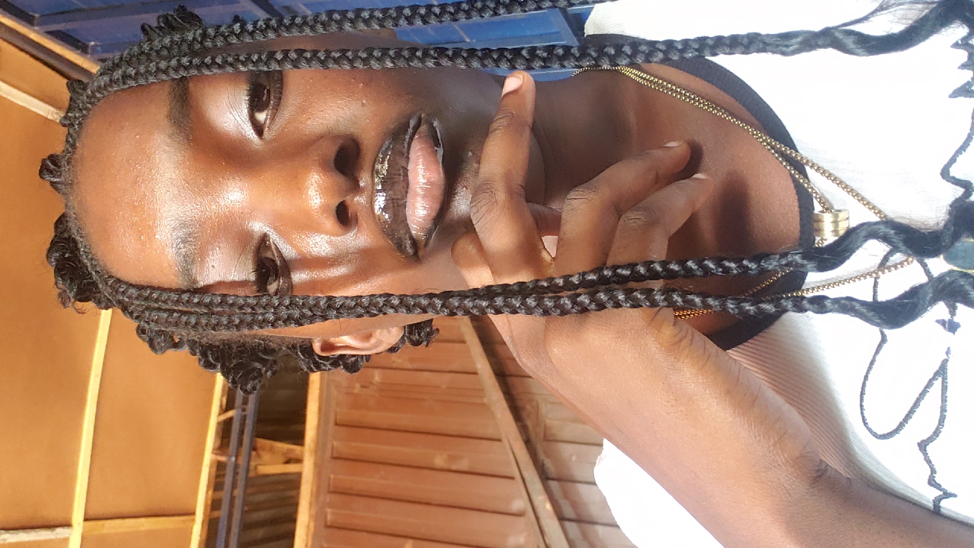 A young Black woman with braided hair, wearing a white shirt and necklaces, poses for a selfie indoors. Her right hand is raised to her chin. The background shows wooden structures and corrugated metal, suggesting an outdoor or semi-outdoor setting, possibly in a workshop or informal structure. The lighting suggests natural light. The location is identified as Kaduna, Nigeria.