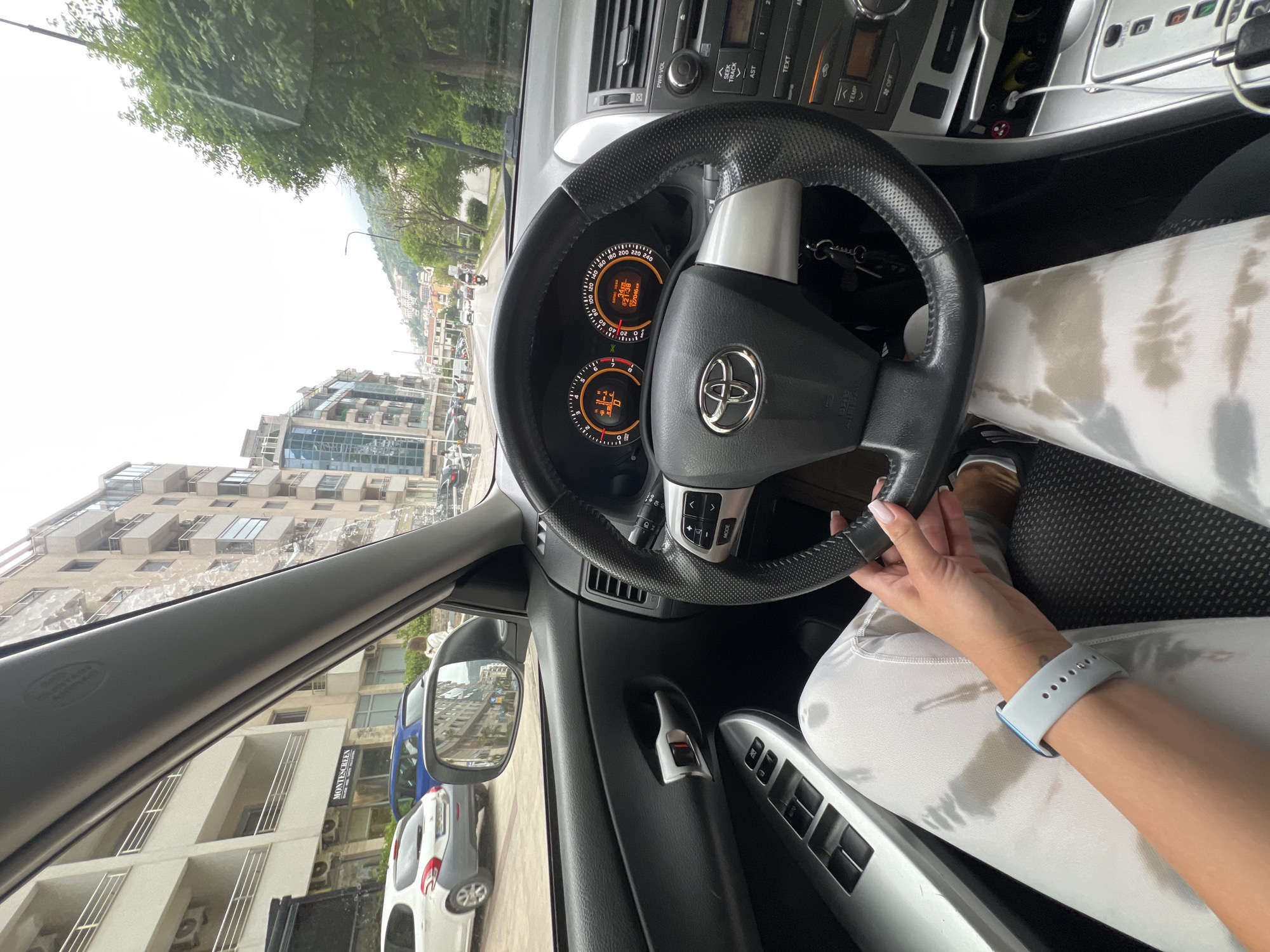 The image captures the interior of a car from a passenger's perspective, focusing on the driver's side and the view outside.
**Subjects:** The primary subject is a person's right arm and hand, gripping the steering wheel of a car. The person is wearing white leggings or pants, which exhibit noticeable dampness or sweat marks, and a light blue smartwatch on their wrist. The car interior includes a black and silver steering wheel with a Toyota logo, a dashboard with an instrument cluster, and parts of the infotainment system and climate controls. Outside the vehicle, multi-story buildings, lush green trees, a street with other cars, and distant hills or mountains are visible.
**Activity or event:** The person appears to be either driving or temporarily stopped in the car, with their hand resting on the steering wheel.
**Notable details:** The scene unfolds on a bright, possibly overcast day, suggesting daytime. The architecture of the buildings outside appears to be European urban style, consistent with the given location context of Budva, Montenegro. The car's instrument cluster shows a digital display with readings like "21136" (potentially mileage) and "1019km". The speedometer is marked up to 240, implying kilometers per hour. The sweat marks on the person's clothing suggest warm weather or recent physical exertion.
**Visible text:**
* On the car's controls and dashboard: "PWR-VOL", "AST", "TEXT", "TEMP", "MODE", "AUTO", and faintly "SIDE AIRBAG".
* On the dashboard display: "21136" and "1019km".
* On the steering wheel: The Toyota logo.
* On a building visible outside the car: "MONTESCREEN".