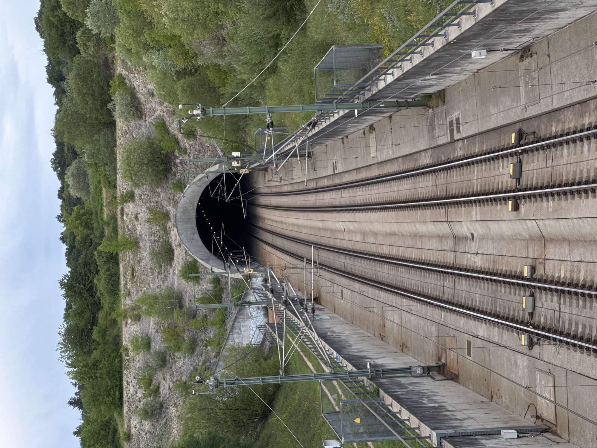 An outdoor, high-angle, vertical shot shows a railway tunnel entrance in Wettstetten, Germany. The tunnel, with a dark, illuminated interior, is carved into a hillside covered with green trees and shrubs. Twin railway tracks run from the foreground to the tunnel entrance, bordered by concrete retaining walls. Overhead power lines, supported by metal poles, extend along the tracks. The sky is overcast and grey.