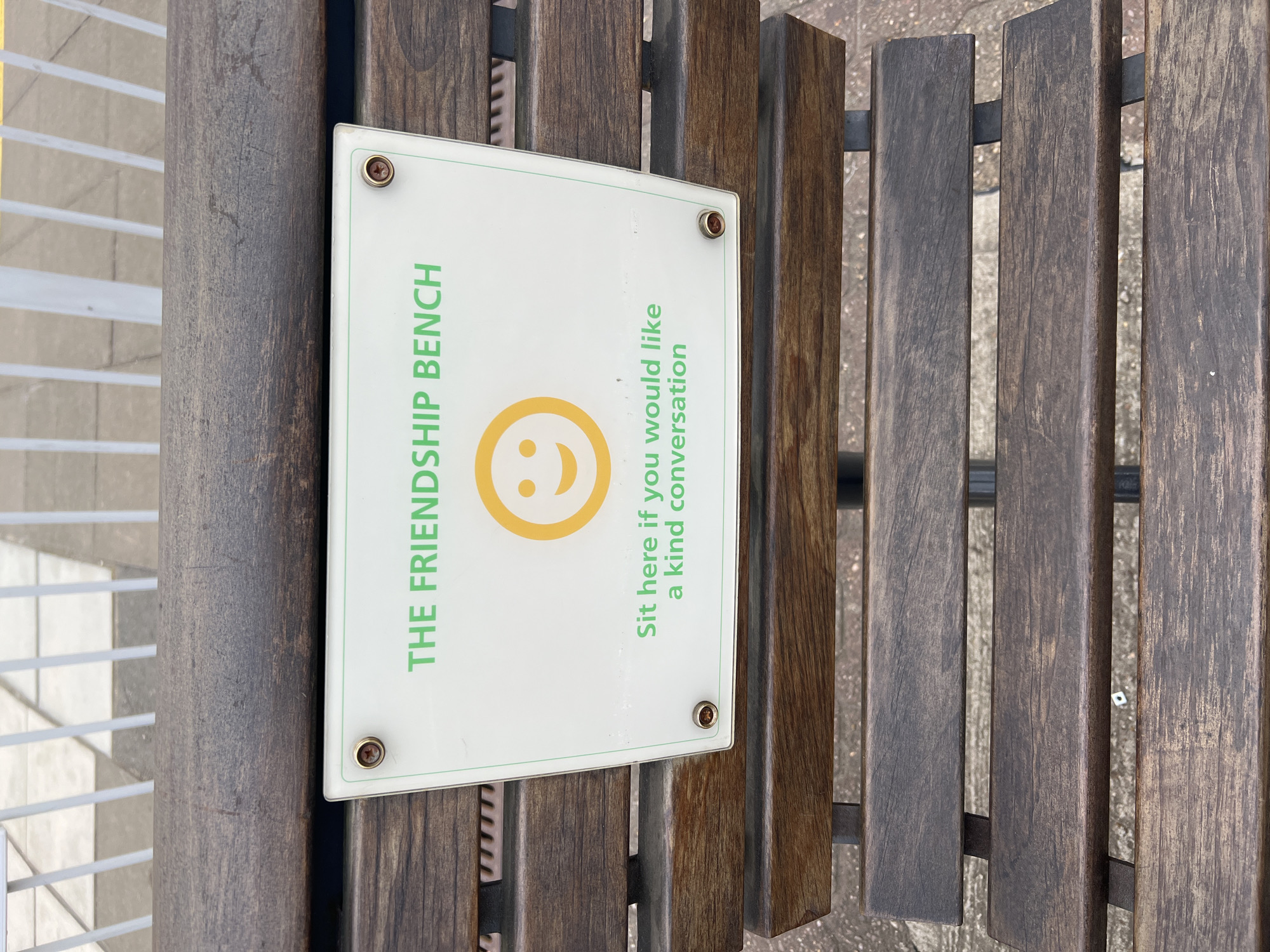 This vertical close-up photograph captures a "Friendship Bench" located in an outdoor public space in London, United Kingdom. The central focus is a white, rectangular sign with rounded corners, prominently displayed on the dark wooden slats of the bench. The sign is affixed with four gold-colored fasteners.
The text on the sign is printed in green. On the left side, oriented vertically, it reads "THE FRIENDSHIP BENCH" in capital letters. A large, yellow smiley face emoticon is positioned in the center of the sign. To the right, also oriented vertically, the text invites: "Sit here if you would like a kind conversation."
The bench is constructed from multiple dark-stained, weathered wooden slats, suggesting a sturdy, utilitarian design typical of public seating. These slats are likely supported by a metal frame, partially visible between the wooden sections. In the background, light-colored paving or concrete ground is visible, indicative of a pedestrian area or urban square. To the far left, a white, grid-like railing or fence can be seen.
No people are present in the image, but the sign's message clearly defines the bench's purpose: to encourage friendly conversations and social interaction, promoting a sense of community and reducing isolation. The scene is illuminated by natural daylight, suggesting it is daytime, possibly under an overcast sky as there are no harsh shadows. The overall impression conveyed is one of warmth and welcome within an urban setting.
