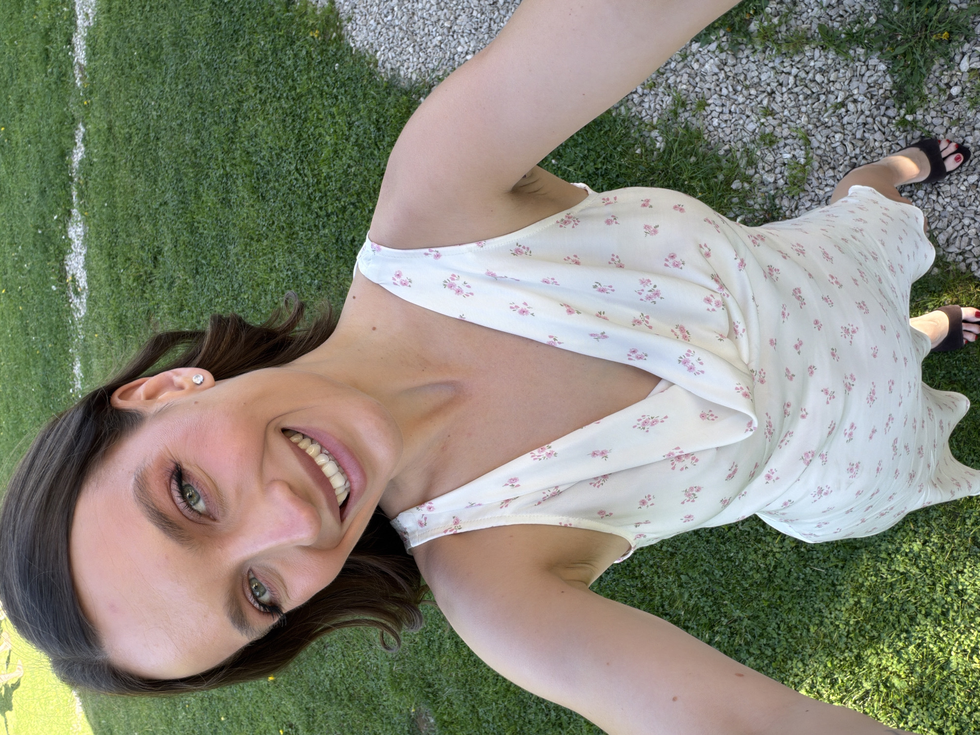 A woman is taking a selfie outdoors. She is wearing a white floral print dress and black sandals with red nail polish. She is smiling and has brown hair and green eyes. The background includes green grass, a white painted line, and gravel. The location is Prudki, Russia.