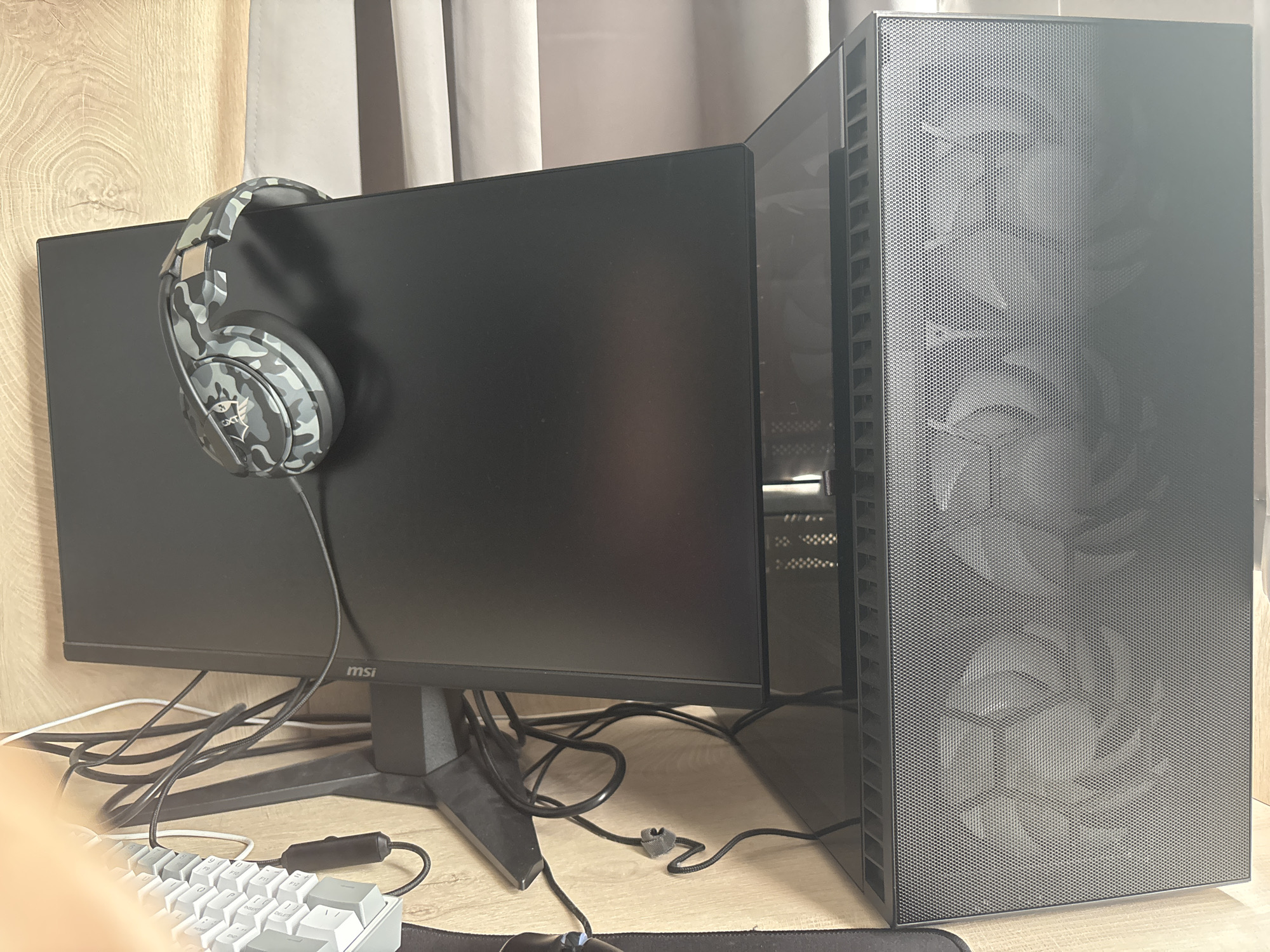 The image displays an indoor computer workstation setup, located in the city of Ruse, Bulgaria. A black computer monitor, with "msi" branding visible on its lower-right bezel, is positioned on a black rectangular stand on a light-colored wooden desk. A pair of circumaural headphones, featuring a grey, black, and white camouflage pattern and a "TRUST" (CXTR) logo on the earcups, is resting on the top edge of the monitor. To the right of the monitor stands a dark grey desktop computer tower, equipped with a perforated mesh front panel that reveals an integrated geometric fan pattern behind it. A reflective side panel of the computer tower is partially visible. Behind and beneath the monitor, a collection of black and white cables are visible, some loosely bundled or tangled, with one black braided cable secured by a small grey clip. In the lower-left foreground, a section of a white mechanical keyboard is visible. The background consists of light-colored, possibly beige or grey, curtains. No individuals are present in the scene, and no explicit actions or interactions are depicted.