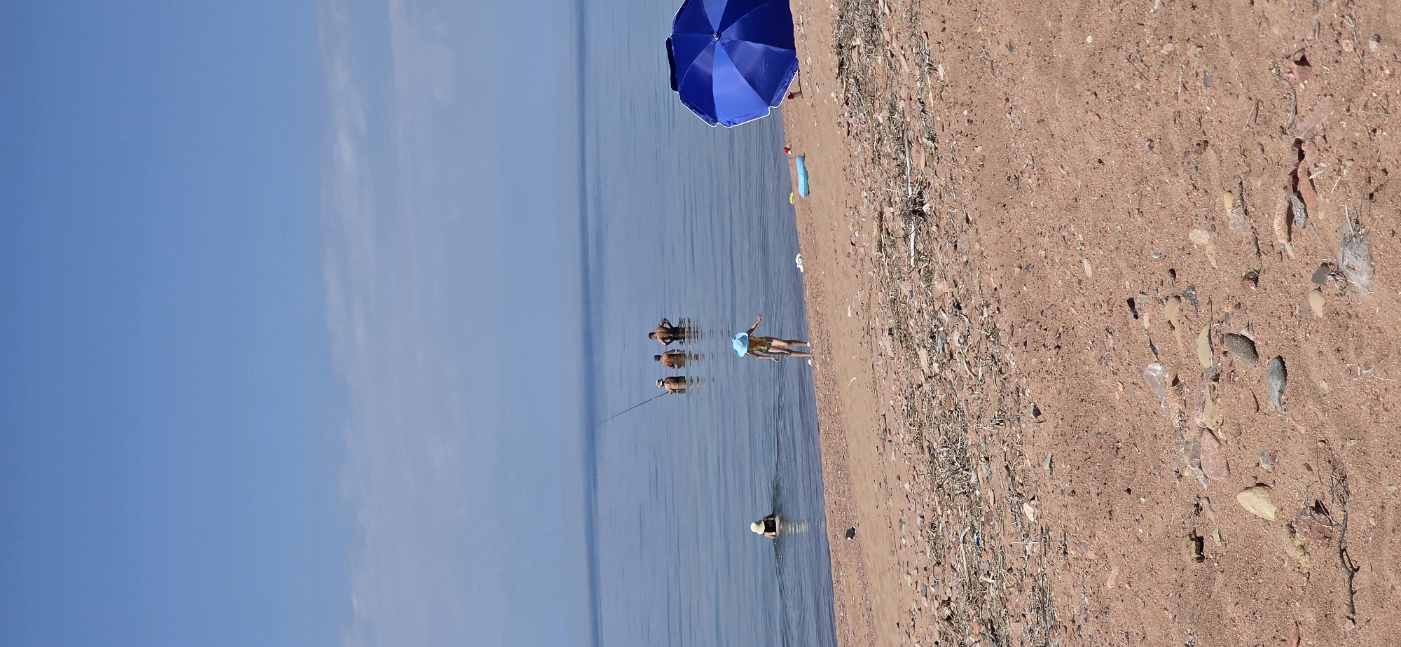 In the city of Tosor, Kyrgyzstan, on a sunny day, several people are enjoying the calm, clear waters of a lake. The beach is composed of reddish sand and small pebbles, with some scattered dry branches and debris. In the foreground, a young child wearing a yellow swimsuit and a blue hat stands near the shore, facing the water. To the right, a large, open blue umbrella provides shade on the sand. Further out in the lake, a person in a straw hat is wading, while three shirtless men are also in the water, with one appearing to be fishing with a long pole. The sky is a bright, clear blue with a few wispy clouds on the horizon, and the water is a tranquil, deep blue.