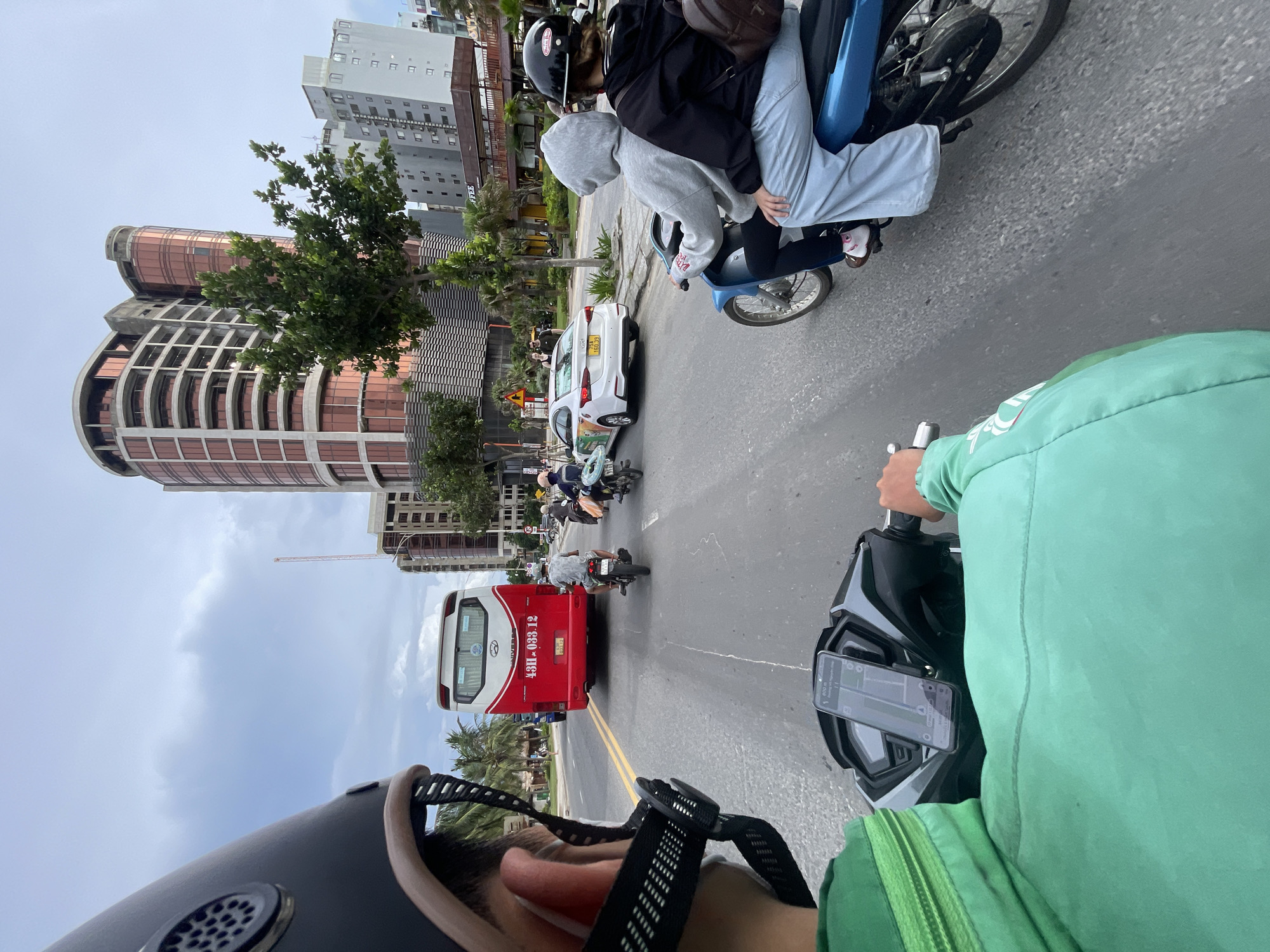 A first-person perspective from a motorcycle shows a street scene in a city. In the foreground, the handlebars of a motorcycle are visible, with a smartphone mounted on them displaying a navigation app. The rider wears a green jacket and a black helmet. Ahead, a red and white bus is traveling in the same direction. To the right of the bus, a white car is moving in the opposite lane. Further down the road, another motorcycle with two passengers is visible. In the background, several tall buildings, some under construction, line the street. The sky is overcast.