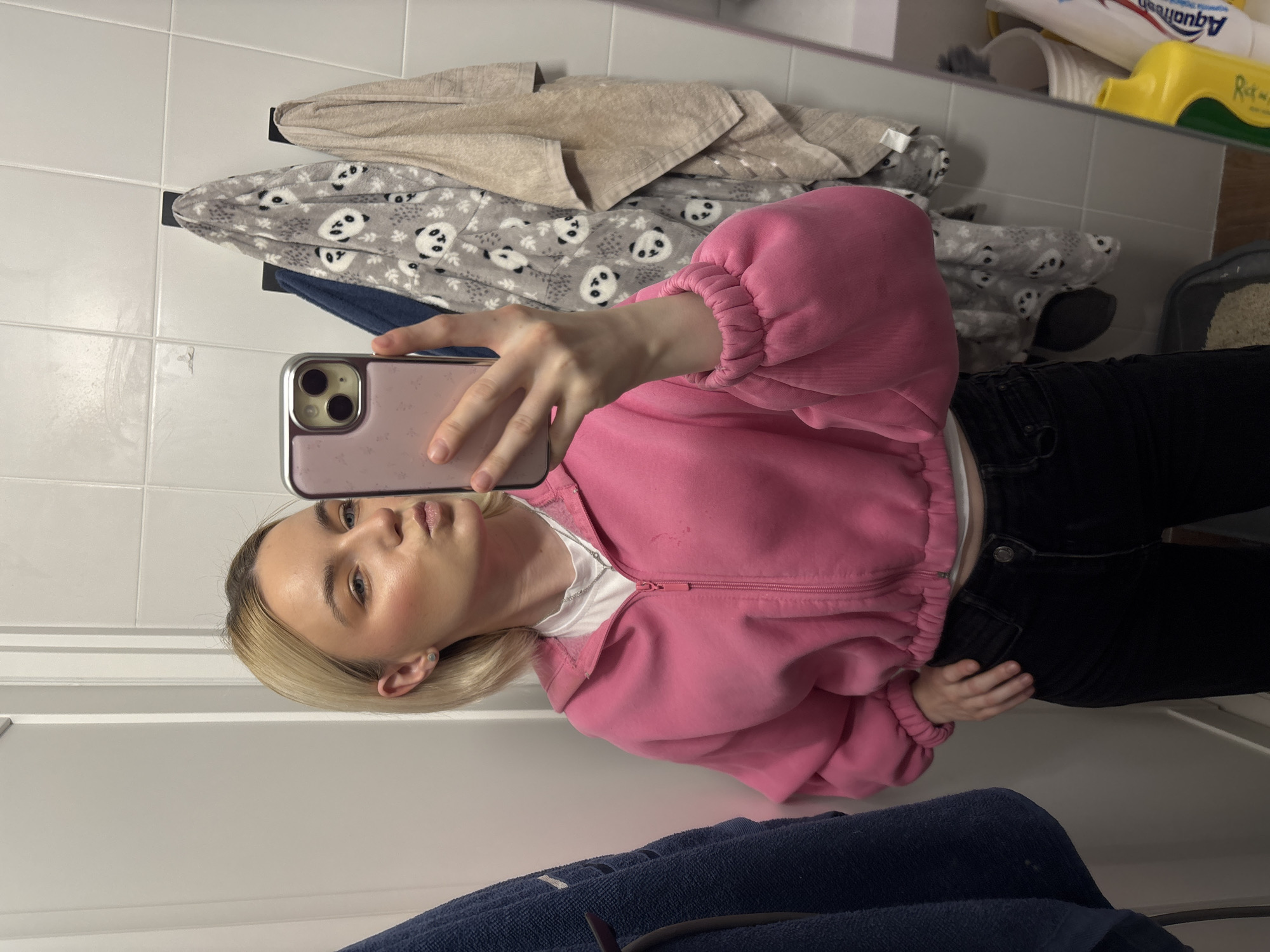 A young person with blonde hair is taking a selfie in what appears to be a bathroom. The person is wearing a pink zip-up sweatshirt and black pants. The background includes white tiled walls with various items hanging on the wall. These items include a light-colored towel, a grey towel with a panda print, and a blue towel. In the upper right corner, there are toiletries and a cat litter box. The photograph was most likely taken at the person's home, but nothing indicates its location in Vsevolozhsk, Russia.