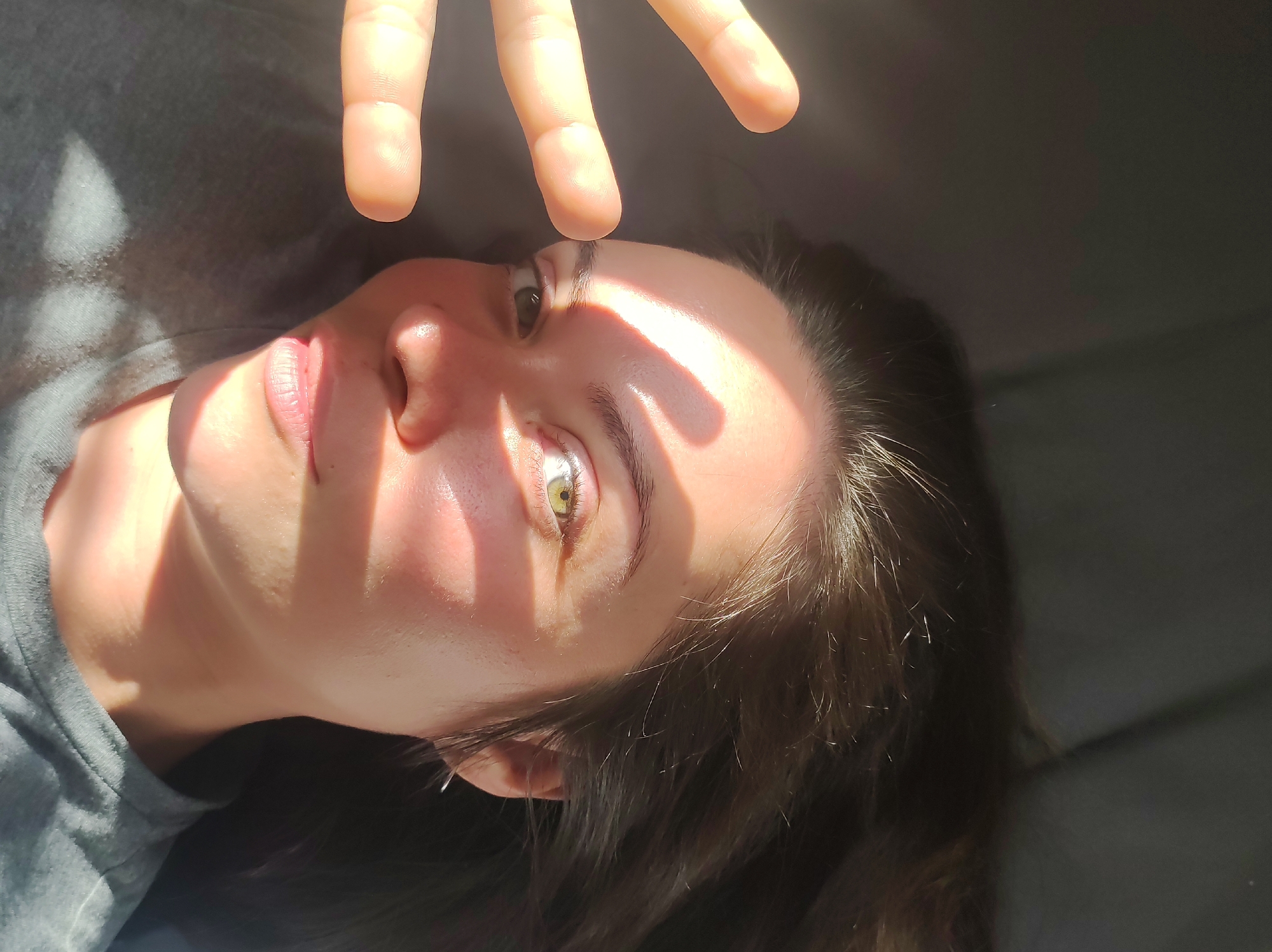 A woman with light skin, brown hair, and green eyes is lying down. Her right hand is raised above her face, creating shadows from the sunlight. She is wearing a gray shirt. The exact location in Chaikovsky, Russia cannot be determined from the image.