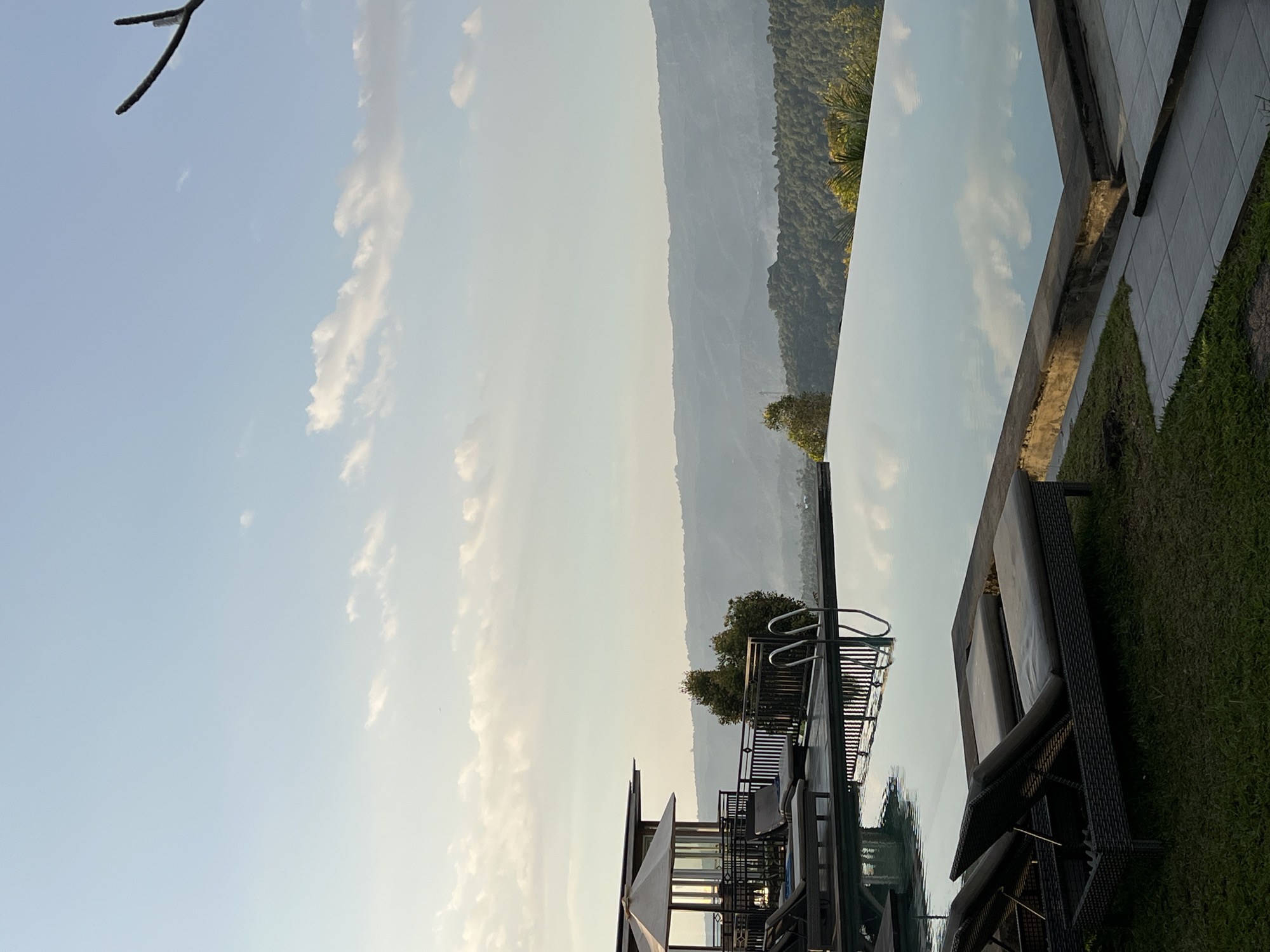The image depicts an infinity pool overlooking a misty valley in Busungbiu, Indonesia. The pool's surface reflects the light blue sky dotted with scattered white clouds. On the left side of the frame, a building with a peaked roof and an umbrella-shaded seating area is visible, along with several empty lounge chairs placed on a wooden deck. Two lounge chairs are prominently featured in the foreground, situated on a patch of green grass. The surrounding landscape is characterized by rolling hills covered in lush green vegetation, fading into a hazy distant horizon. A tree branch extends into the upper right corner of the image.