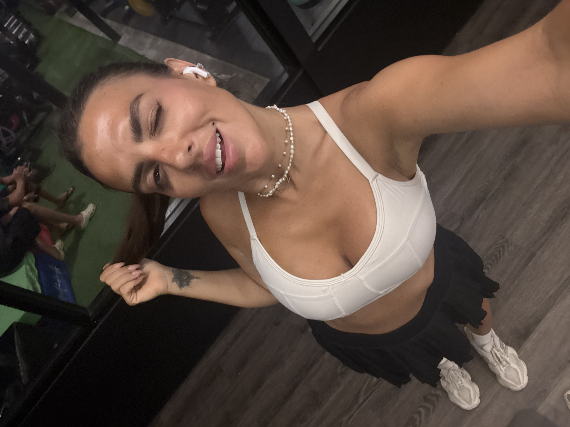 A woman is in a gym in North Kuta, Indonesia. She is wearing a white sports bra, a black tennis skirt, and white sneakers. She has a pearl necklace and a white earbud in her ear. She has brown hair pulled back into a ponytail and a tattoo on her left wrist. The background shows gym equipment and wooden floors. She is winking and taking a selfie.